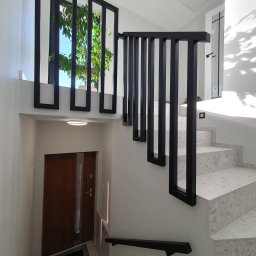 SpawPro - Nowoczesne schody wewnętrzne z szarymi stopniami, czarną balustradą z pionowych elementów i oknem z widokiem na zieleń, oświetlone światłem słonecznym wpadającym przez okno i lampami sufitowymi.