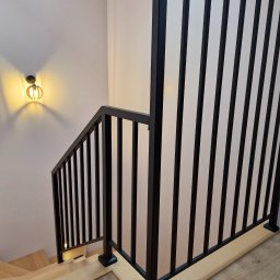 SpawPro - Nowoczesne schody z drewnianymi stopniami i czarną, metalową balustradą z pionowymi szczeblami, oświetlone dekoracyjną lampą ścienną.