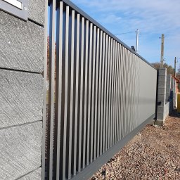 SpawPro - Nowoczesna, przesuwna brama wjazdowa z pionowych profili, zamontowana na słupkach z szarego betonu architektonicznego, na tle żwirowego podjazdu i błękitnego nieba.