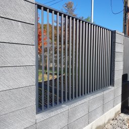 SpawPro - Nowoczesne ogrodzenie panelowe z betonowych bloczków i metalowych sztachet w kolorze grafitowym, widok z zewnątrz na tle błękitnego nieba i jesiennych drzew.