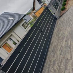 SpawPro - Nowoczesna, czarna brama wjazdowa z poziomymi szczeblami, widoczna z perspektywy chodnika z szarej kostki brukowej, na tle domu z drewnianymi drzwiami.