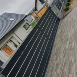 Nowoczesna, czarna brama wjazdowa z poziomymi szczeblami, widoczna z perspektywy chodnika z szarej kostki brukowej, na tle domu z drewnianymi drzwiami.