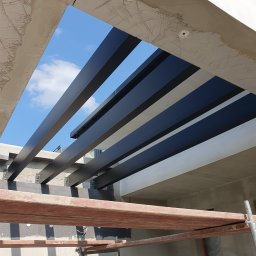 SpawPro - Widok z dołu na konstrukcję z ciemnych, metalowych profili tworzących zadaszenie, z fragmentem nieba widocznym pomiędzy nimi, oraz rusztowaniem w dolnej części kadru.