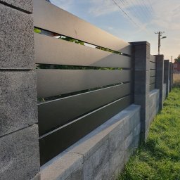 SpawPro - Nowoczesne ogrodzenie z poziomych, grafitowych paneli metalowych, osadzone na betonowej podmurówce z szarej kostki, widok z boku, w tle trawa i słup energetyczny.