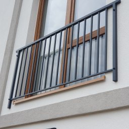 SpawPro - Nowoczesna, grafitowa balustrada balkonowa z pionowymi prętami, zamontowana na elewacji budynku z drewnianą ramą okienną w tle.