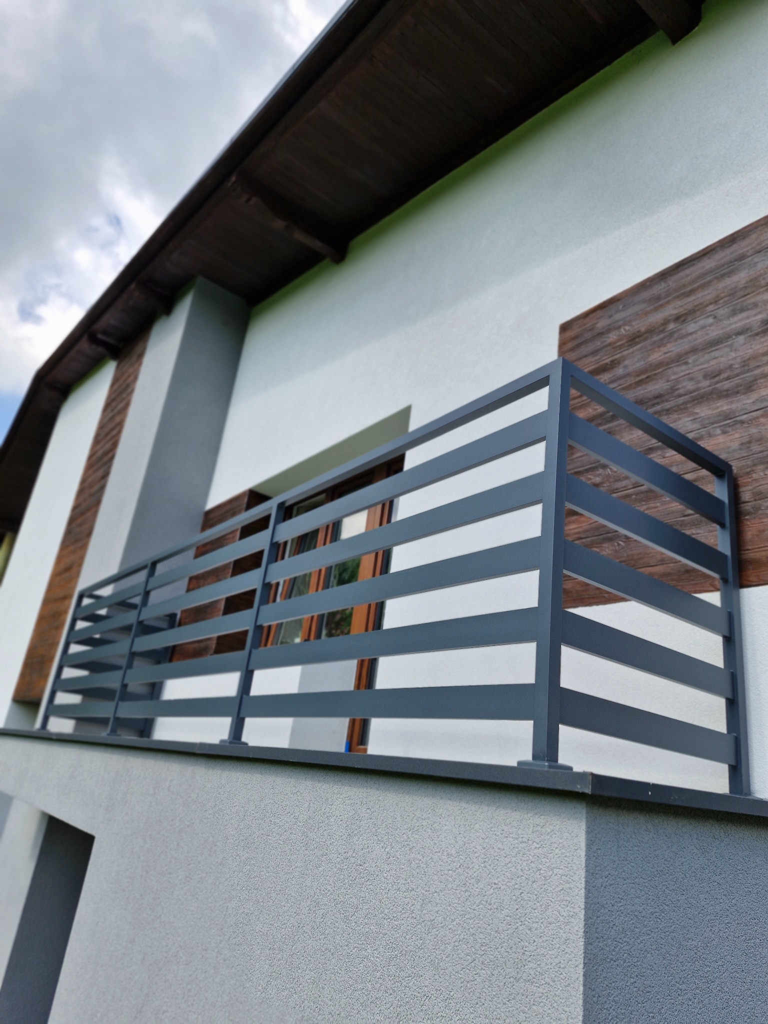 Nowoczesna, szara balustrada balkonowa z poziomych profili na tle jasnej elewacji budynku z drewnianymi elementami dekoracyjnymi.