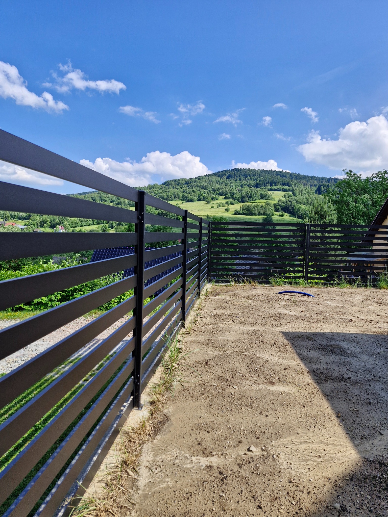Nowoczesne, poziome ogrodzenie panelowe w kolorze antracytowym, z widokiem na pagórkowaty krajobraz i błękitne niebo z chmurami.