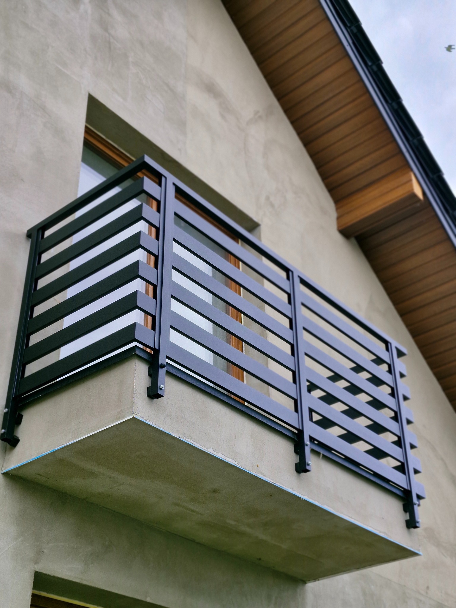 Nowoczesna, czarna balustrada balkonowa z poziomych profili, zamontowana na surowej elewacji budynku z drewnianą podbitką dachu.