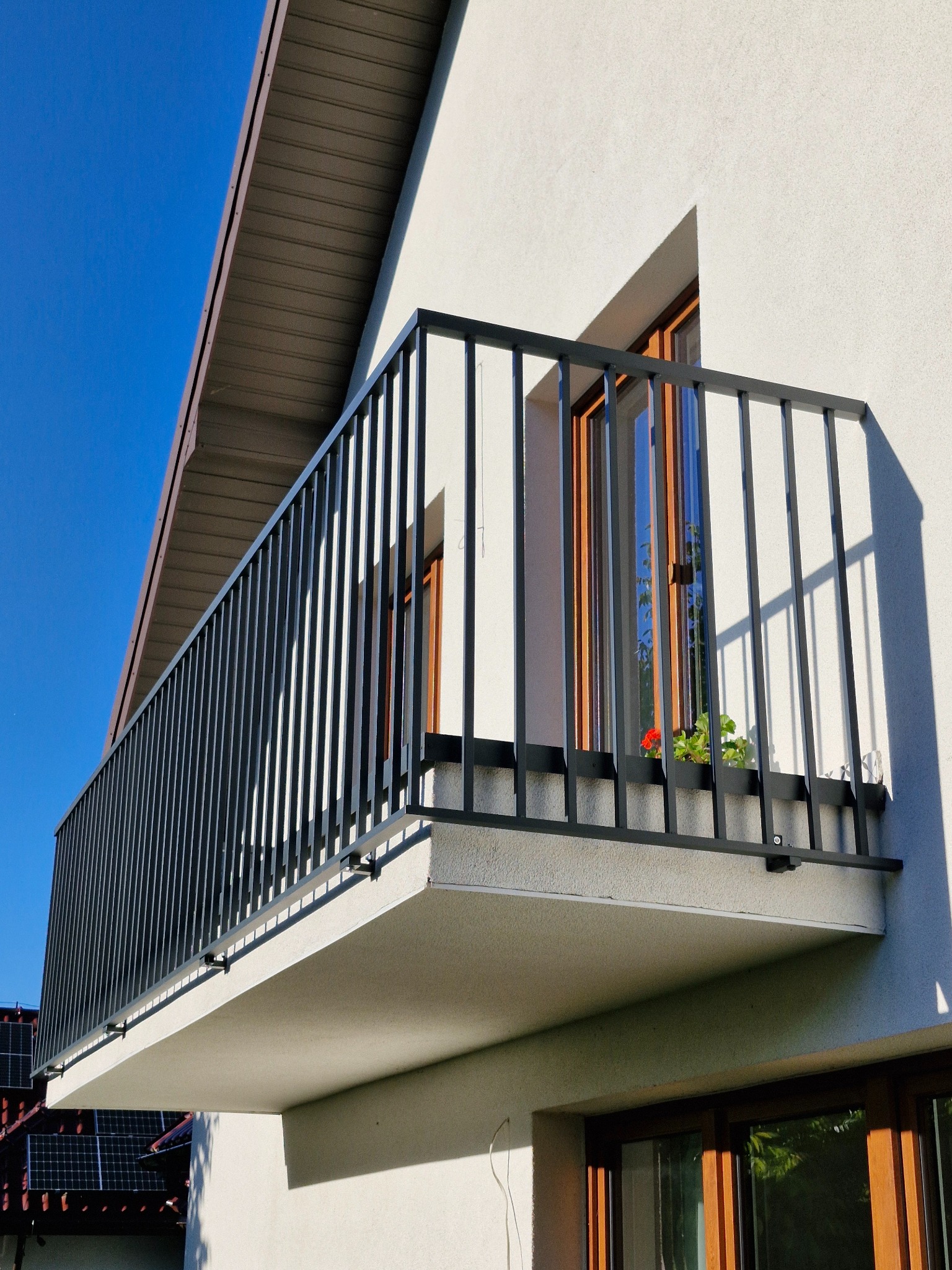 Nowoczesna balustrada balkonowa z pionowych, czarnych prętów, zamontowana do białego balkonu budynku z oknami o drewnianych ramach; w tle widoczne panele słoneczne na dachu.