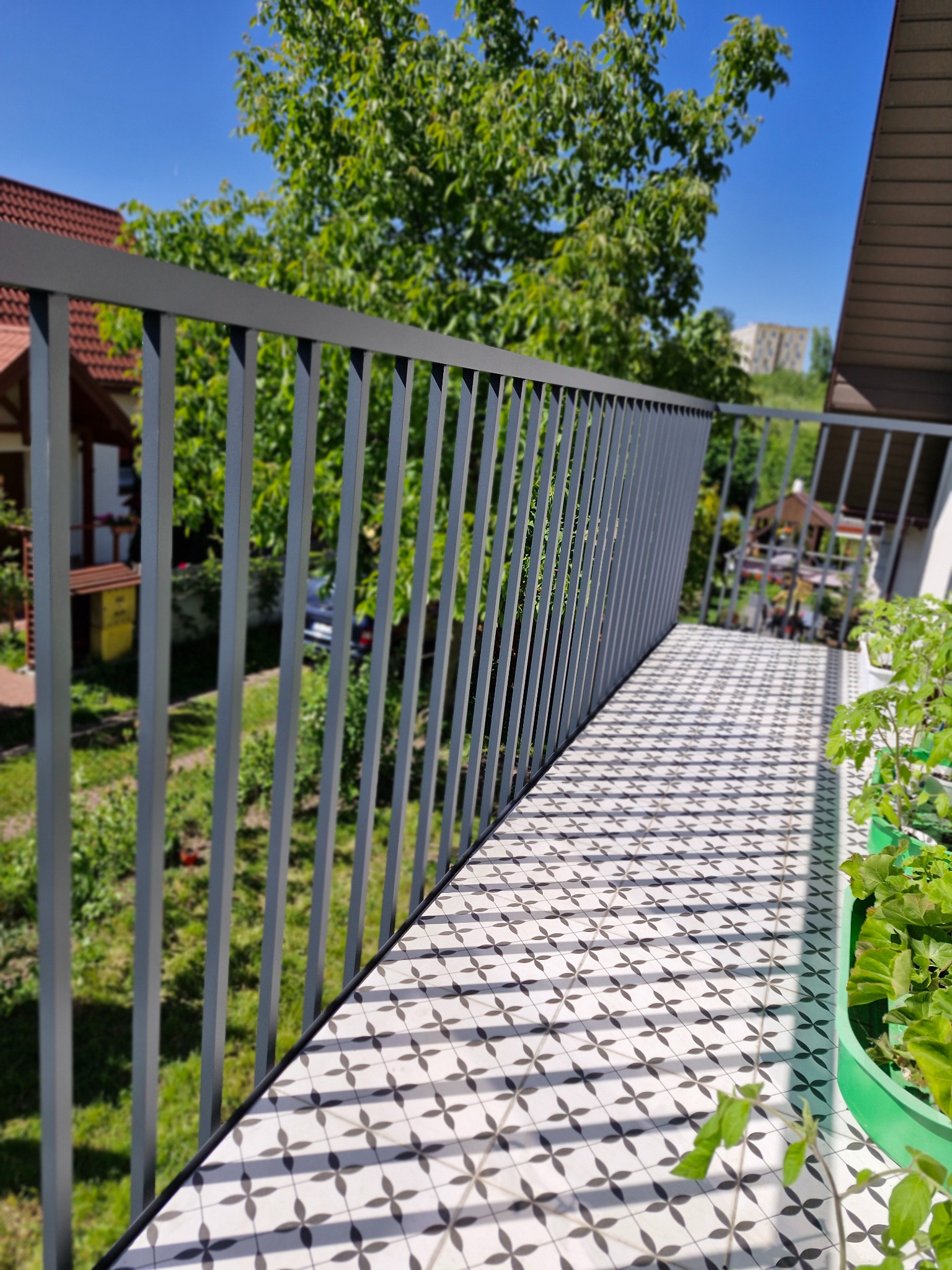 Nowoczesna, szara balustrada balkonowa z pionowych profili, wzorzysta podłoga balkonu w biało-czarne kwiaty, widok na zieleń ogrodu i zabudowania w tle, donice z sadzonkami z prawej strony.