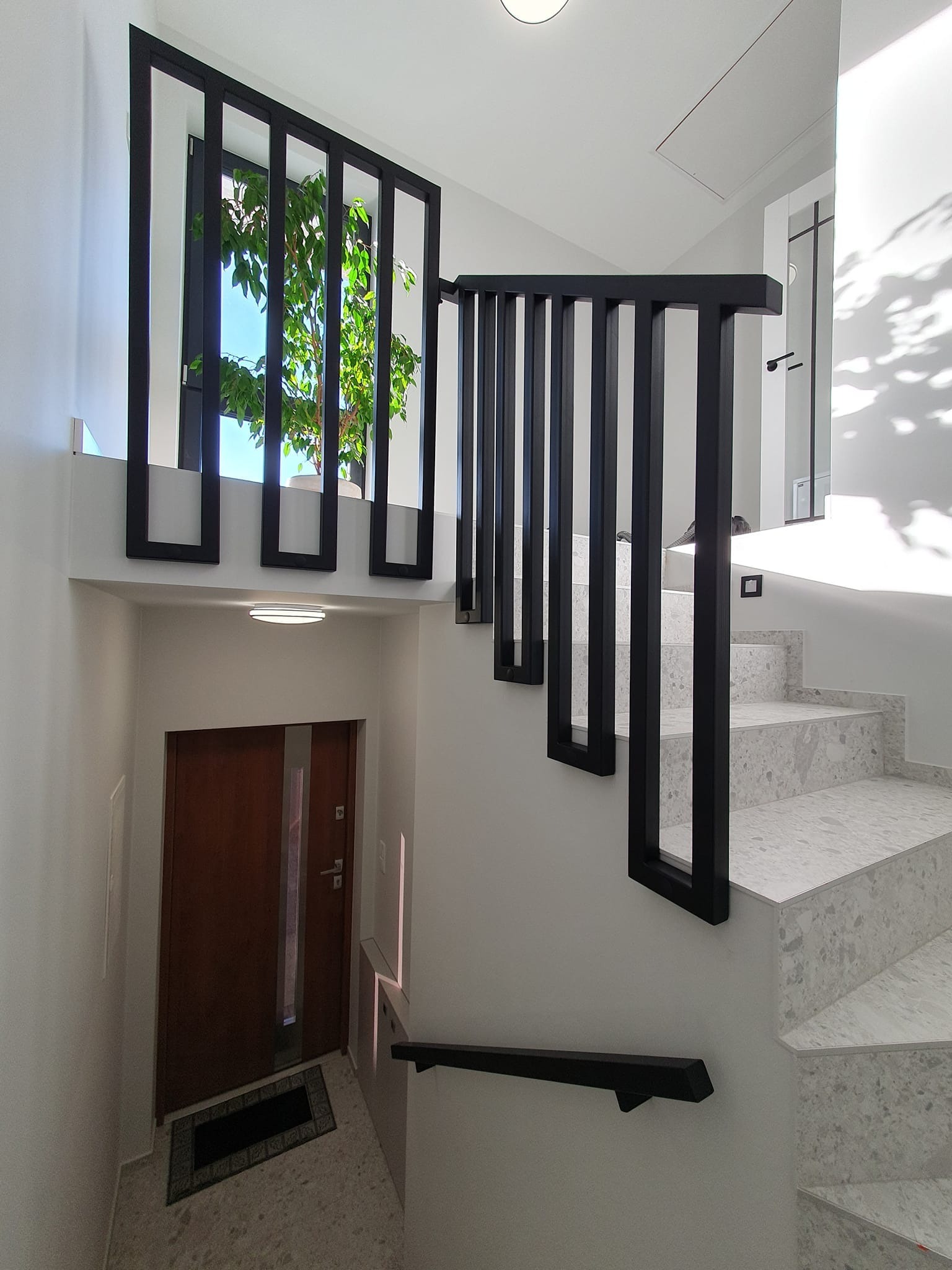 Nowoczesne schody wewnętrzne z szarymi stopniami, czarną balustradą z pionowych elementów i oknem z widokiem na zieleń, oświetlone światłem słonecznym wpadającym przez okno i lampami sufitowymi.