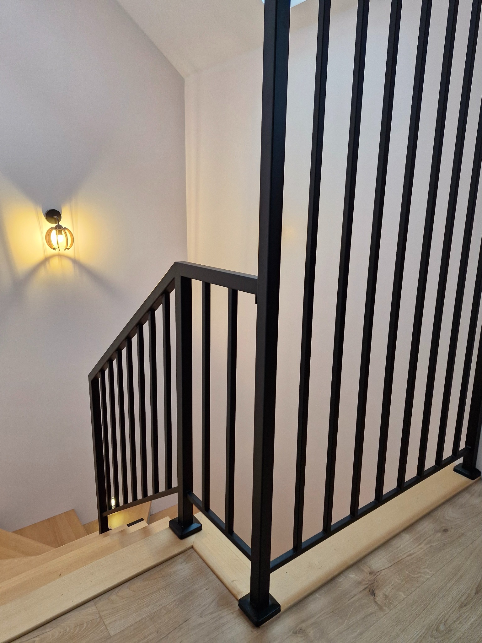 Nowoczesne schody z drewnianymi stopniami i czarną, metalową balustradą z pionowymi szczeblami, oświetlone dekoracyjną lampą ścienną.