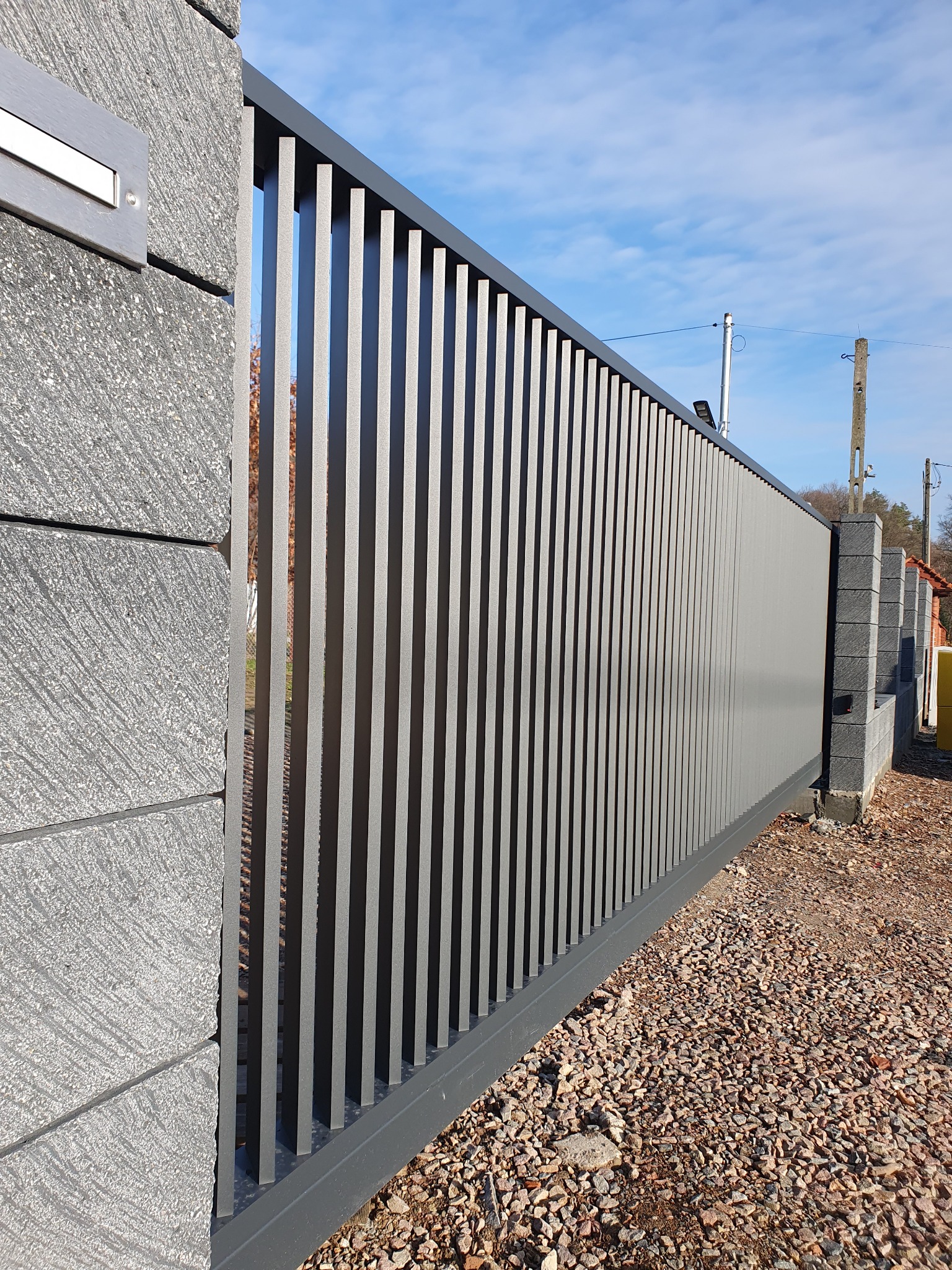 Nowoczesna, przesuwna brama wjazdowa z pionowych profili, zamontowana na słupkach z szarego betonu architektonicznego, na tle żwirowego podjazdu i błękitnego nieba.
