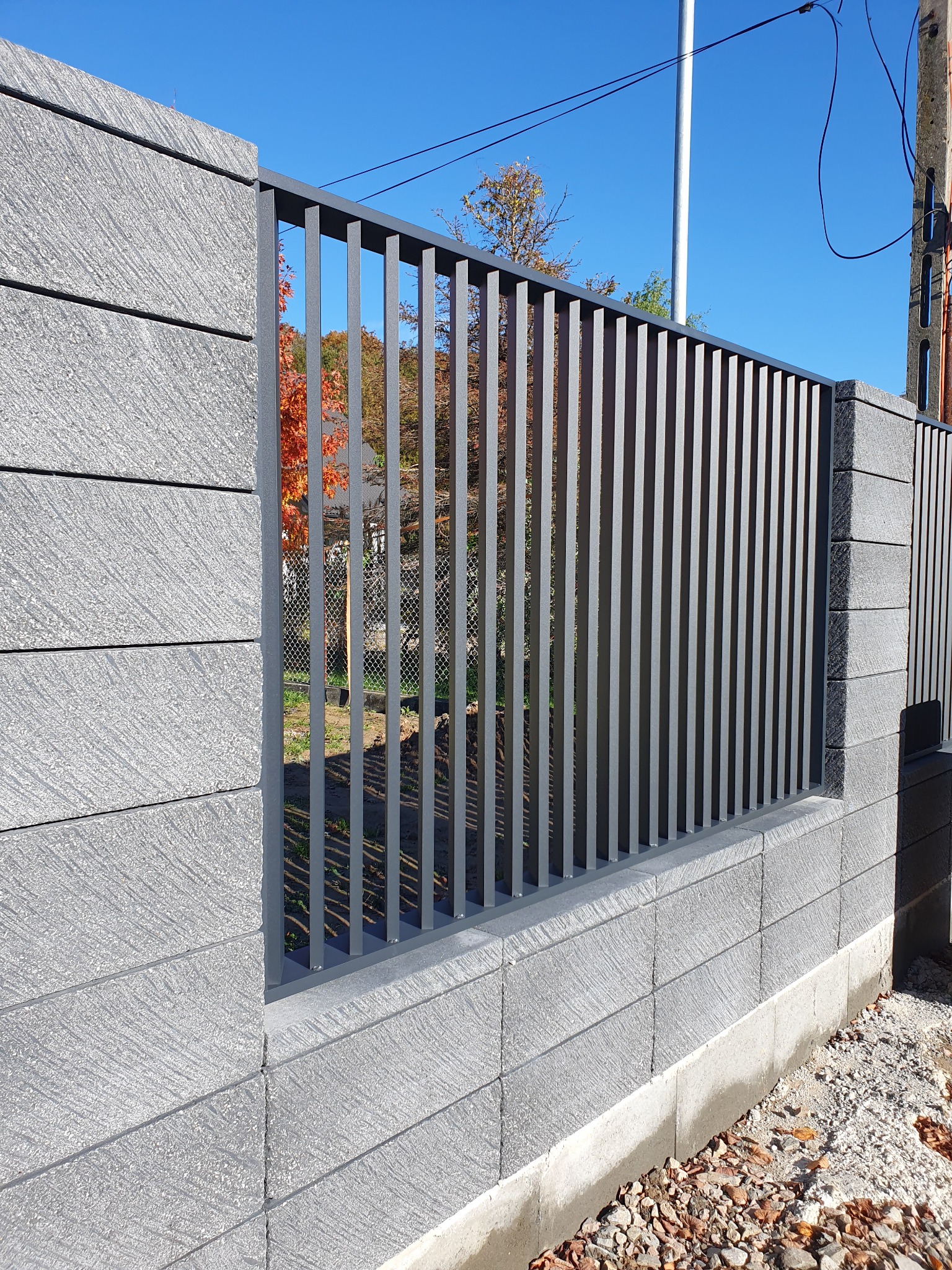 Nowoczesne ogrodzenie panelowe z betonowych bloczków i metalowych sztachet w kolorze grafitowym, widok z zewnątrz na tle błękitnego nieba i jesiennych drzew.