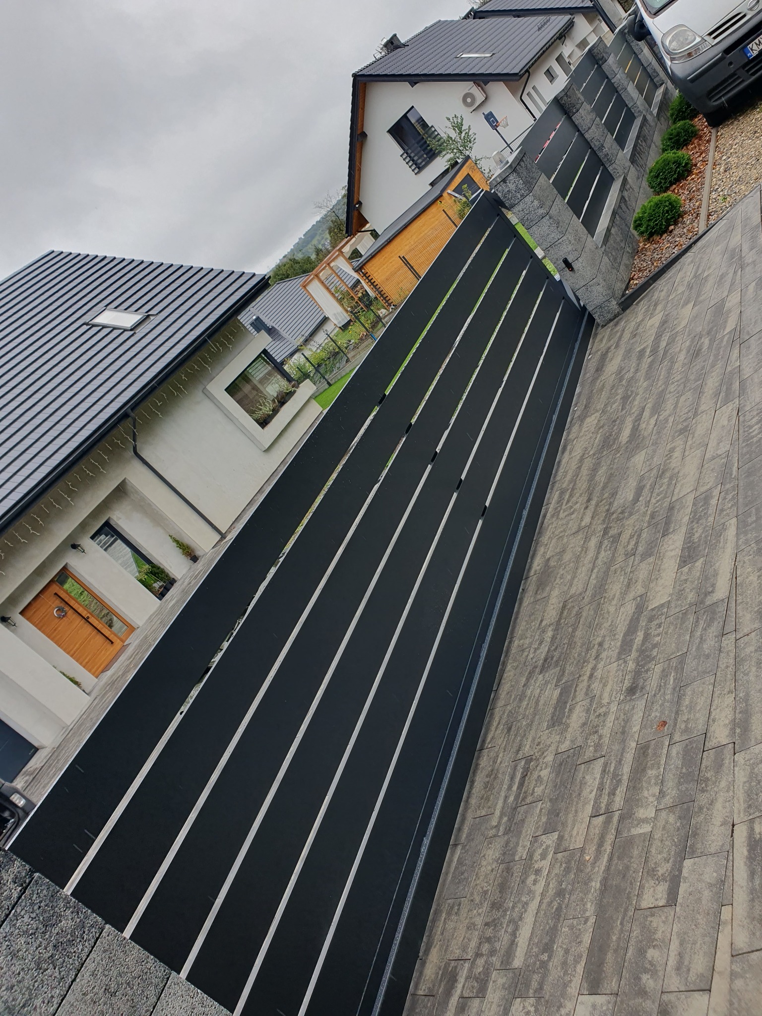 Nowoczesna, czarna brama wjazdowa z poziomymi szczeblami, widoczna z perspektywy chodnika z szarej kostki brukowej, na tle domu z drewnianymi drzwiami.