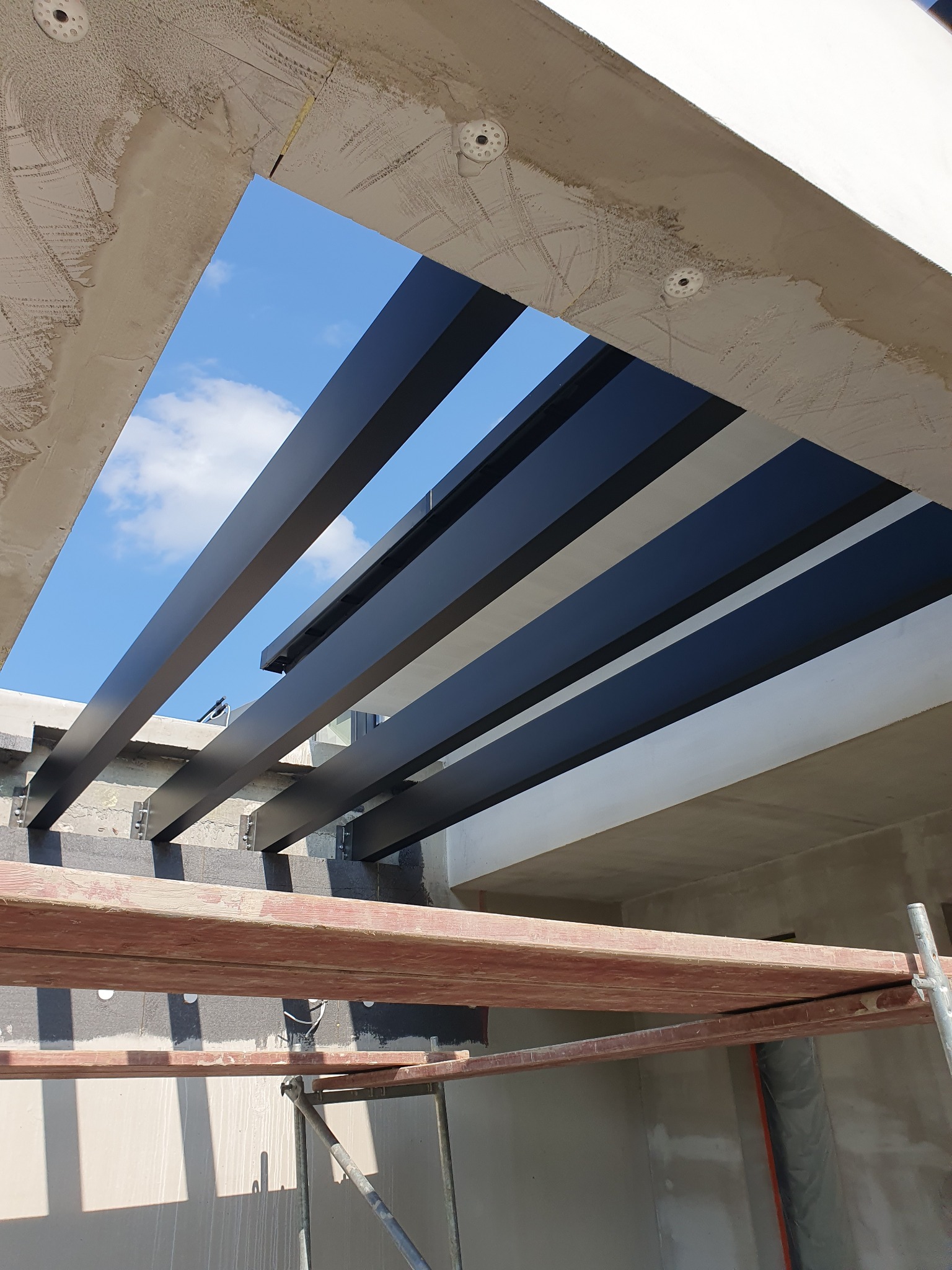 Widok z dołu na konstrukcję z ciemnych, metalowych profili tworzących zadaszenie, z fragmentem nieba widocznym pomiędzy nimi, oraz rusztowaniem w dolnej części kadru.