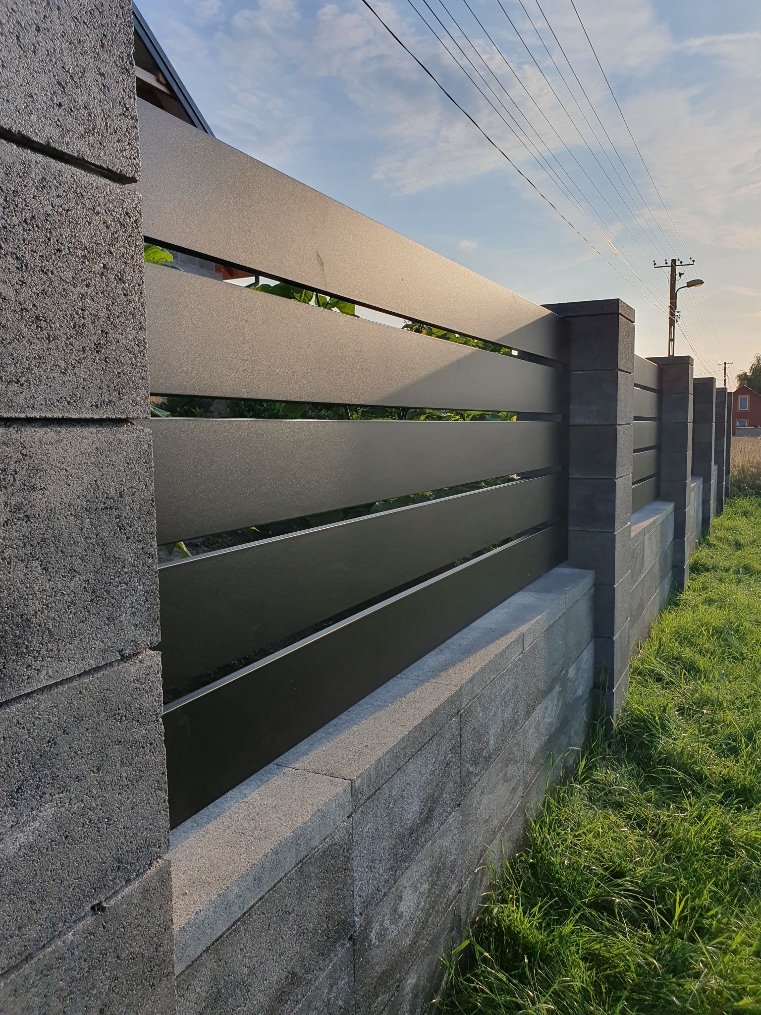 Nowoczesne ogrodzenie z poziomych, grafitowych paneli metalowych, osadzone na betonowej podmurówce z szarej kostki, widok z boku, w tle trawa i słup energetyczny.