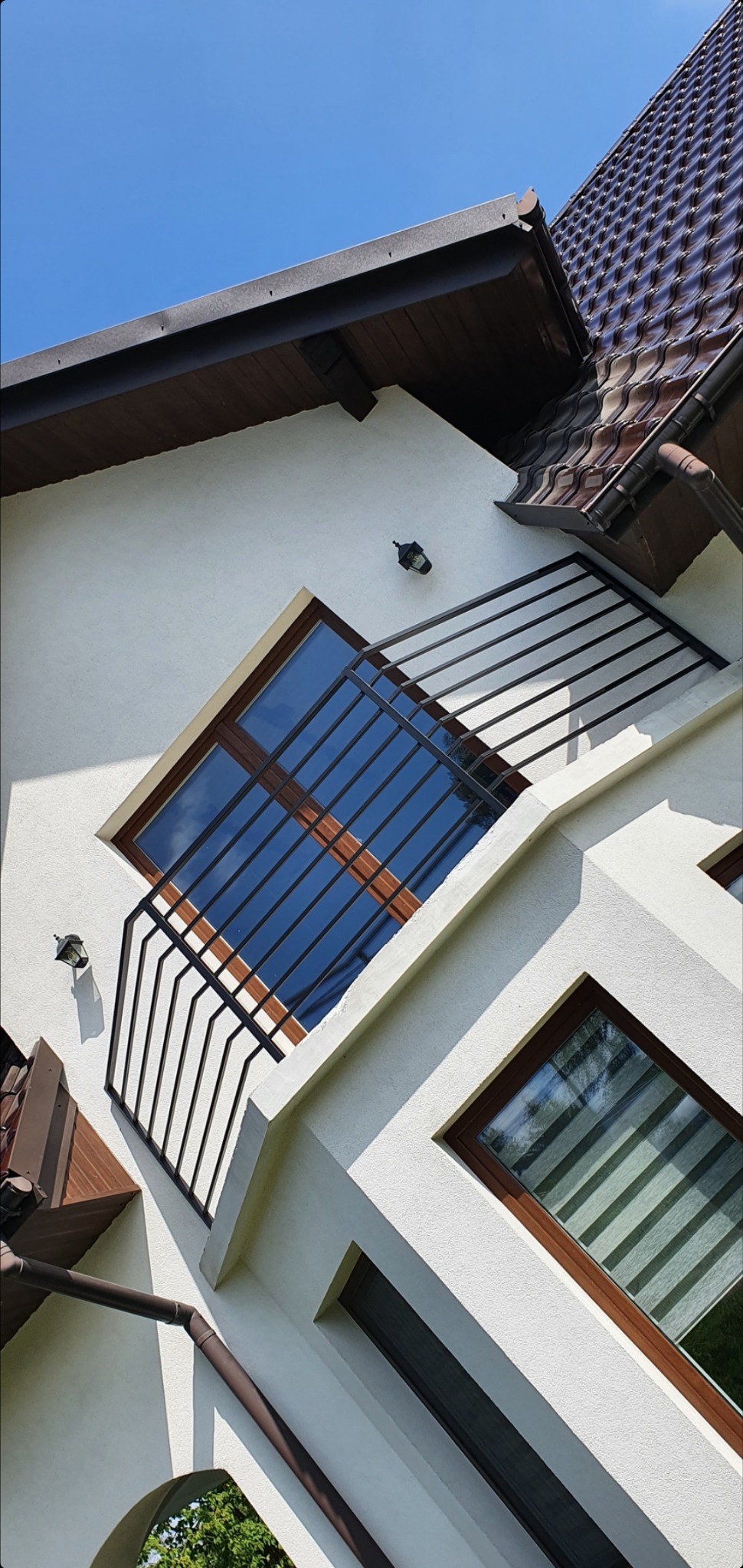 Nowoczesna balustrada balkonowa z poziomych, czarnych prętów na białym budynku z brązowymi oknami i dachem w słoneczny dzień.