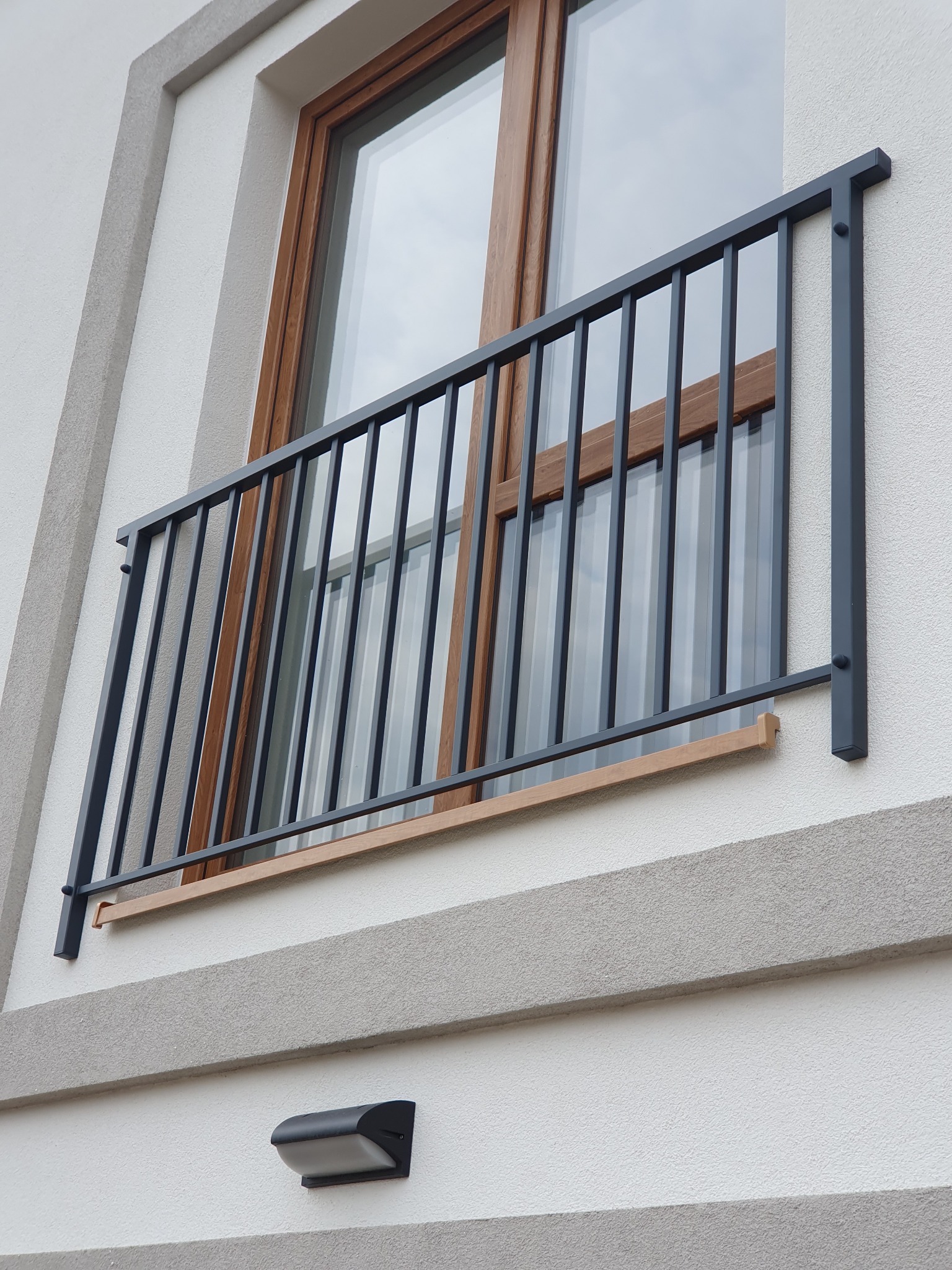 Nowoczesna, grafitowa balustrada balkonowa z pionowymi prętami, zamontowana na elewacji budynku z drewnianą ramą okienną w tle.
