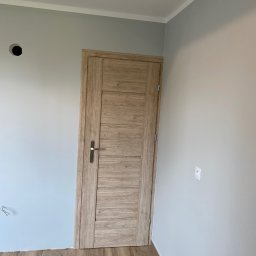Arkadiusz Rech - Wnętrze pokoju po remoncie: drewnopodobne panele podłogowe, pomalowane na szaro ściany z widocznym gniazdkiem elektrycznym i otworem na instalację, zamontowane drzwi wewnętrzne z drewnianą okleiną...
