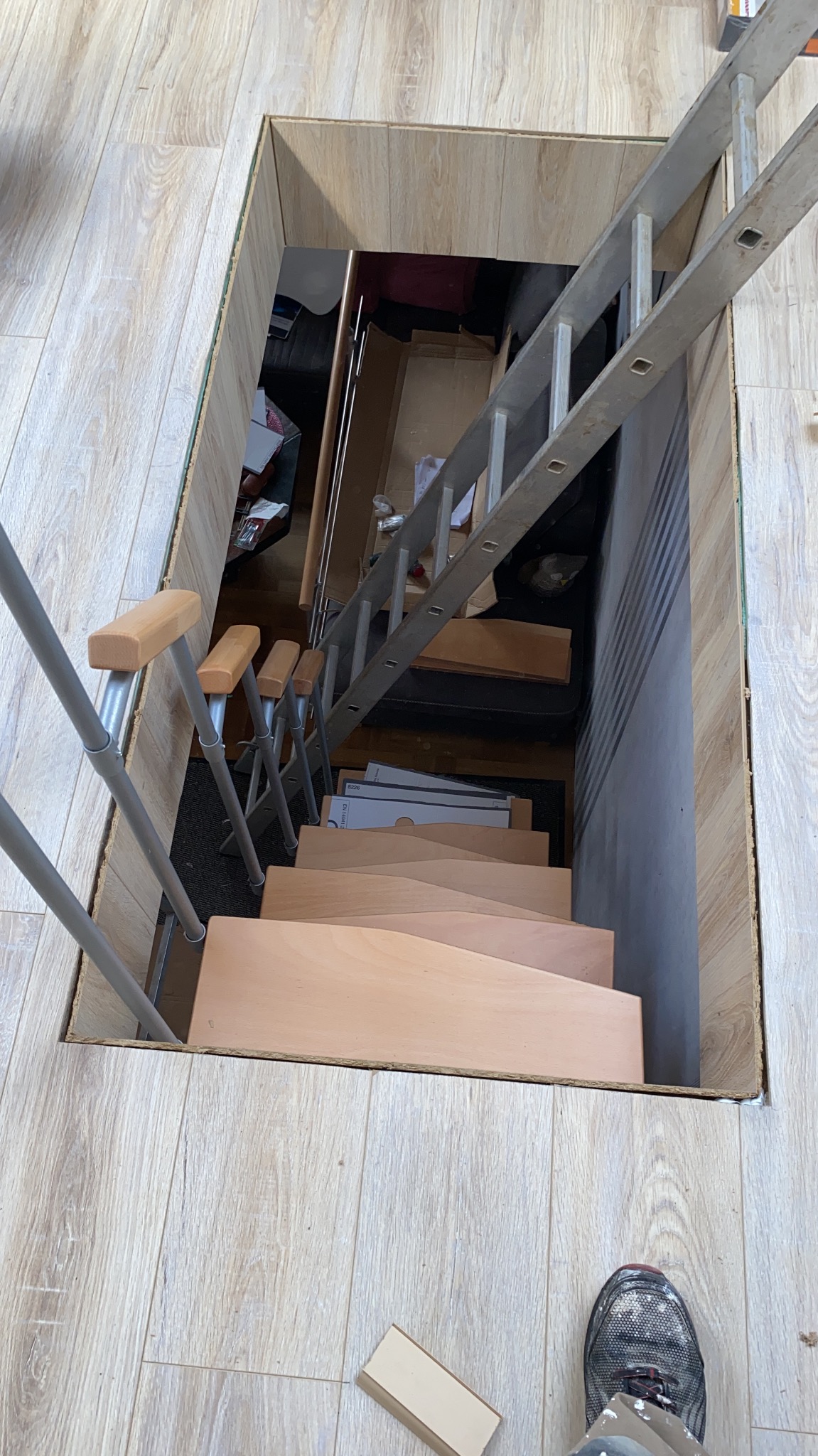 Wysokie ujęcie na schody prowadzące do piwnicy, z widocznymi panelami podłogowymi dookoła otworu, fragment drabiny oparty o ścianę, oraz stopa w roboczym obuwiu w prawym dolnym rogu.