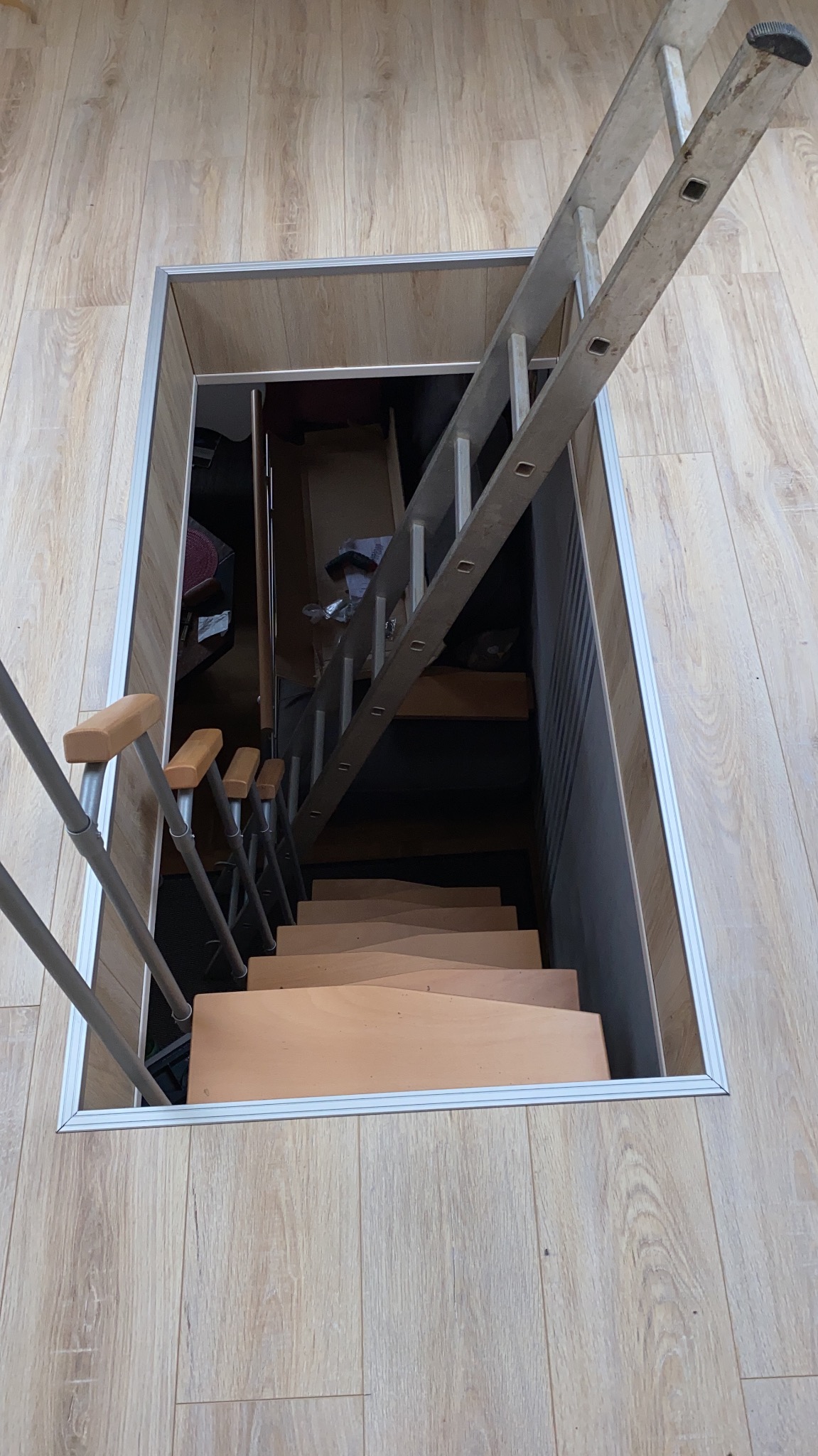 Widok z góry na otwór w podłodze wykończonej panelami, prowadzący do poddasza z drewnianymi schodami i rozkładaną metalową drabinką.