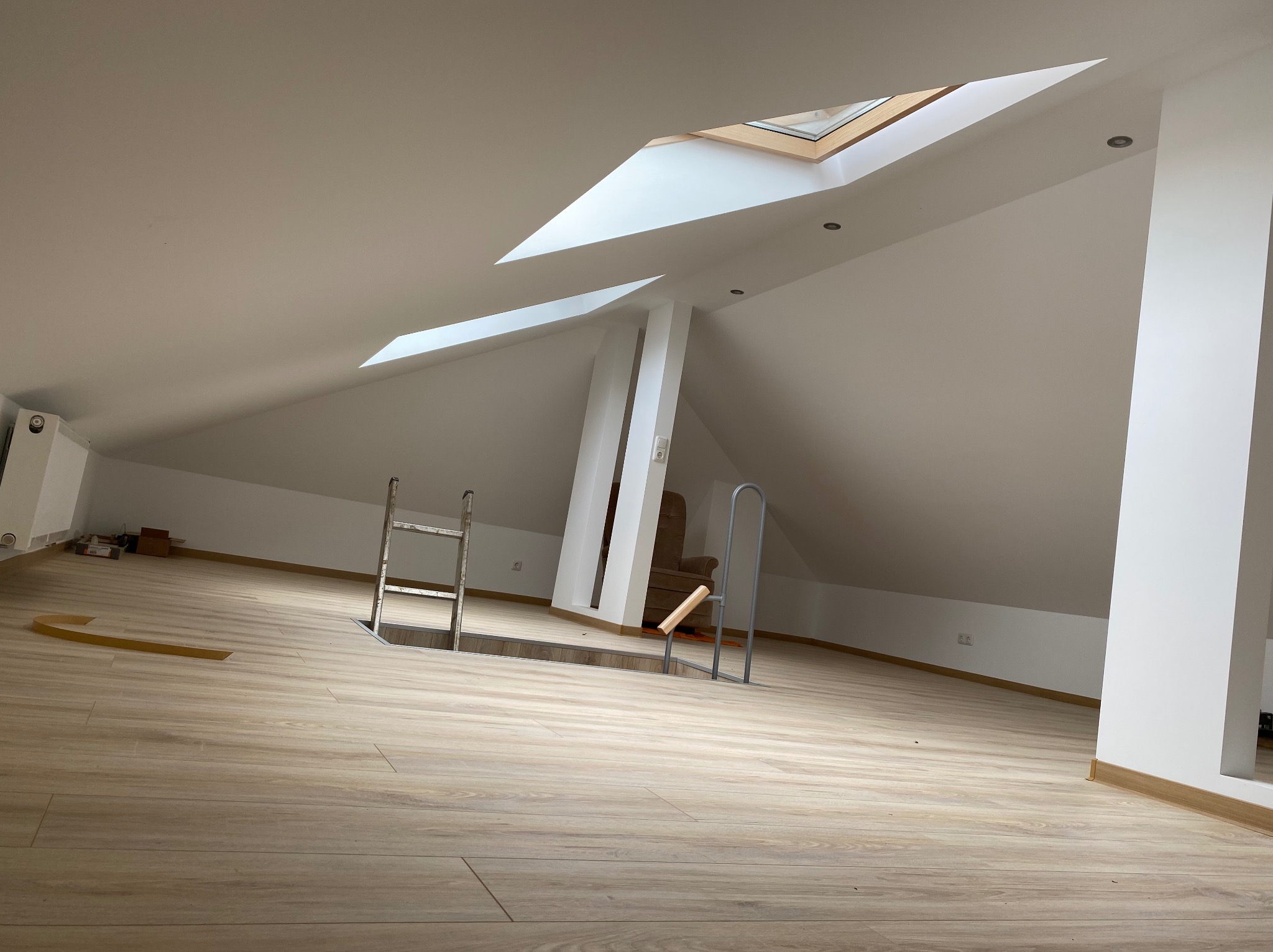 Poddasze po remoncie z drewnopodobnymi panelami podłogowymi, schodami strychowymi i oknem dachowym, widok na fragment fotela w tle.