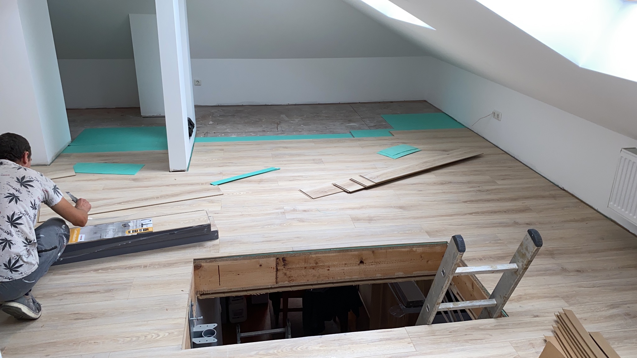 Montaż paneli podłogowych w skosach poddasza, widoczny pracownik przycinający panel, rozłożone podkłady izolacyjne, niezabezpieczony otwór w podłodze z drabiną.