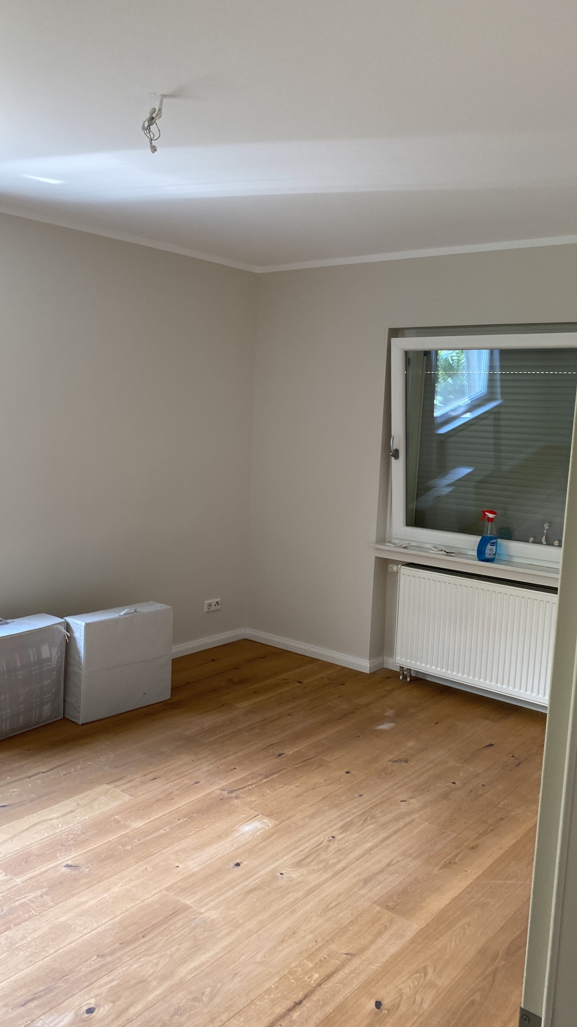 Pusty pokój po remoncie z drewnianą podłogą, grzejnikiem pod oknem i przygotowanymi panelami w opakowaniach, widoczne kable wystające z sufitu.