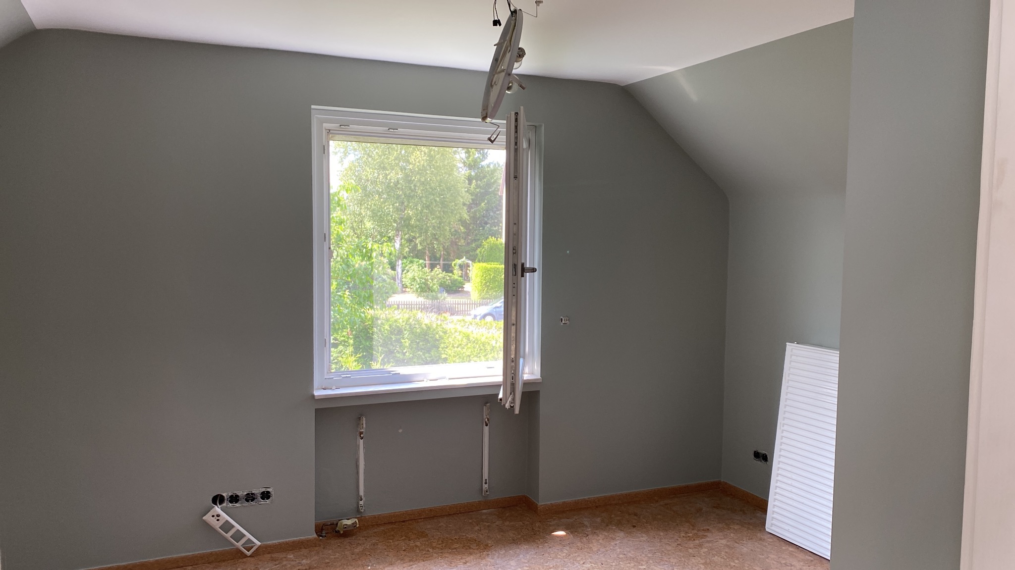 Pokój w trakcie prac wykończeniowych, ściany pomalowane na szaro, widoczne wyprowadzenia elektryczne, grzejnik oraz niedokończona podłoga.