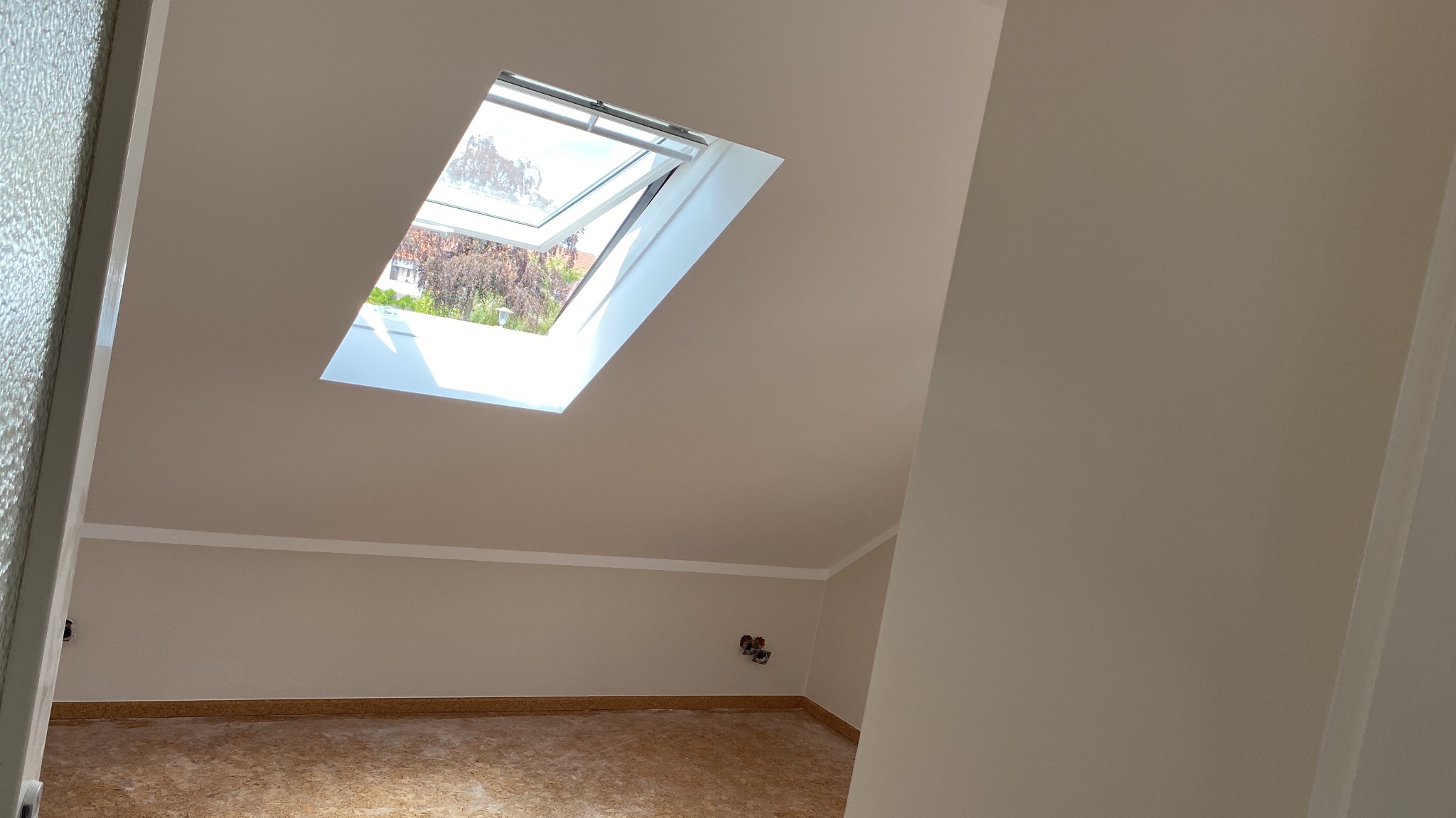 Poddasze po remoncie z oknem dachowym, widoczne świeżo pomalowane ściany w odcieniach beżu, surowe wykończenie podłogi i wystające kable elektryczne.