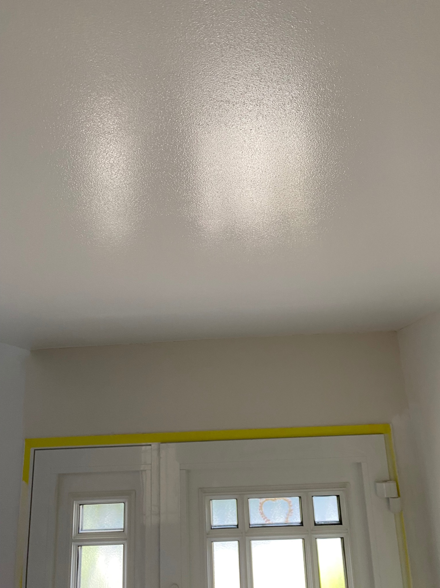 Świeżo pomalowany, biały sufit z widoczną fakturą farby, żółta taśma malarska zabezpiecza ramę okienną.
