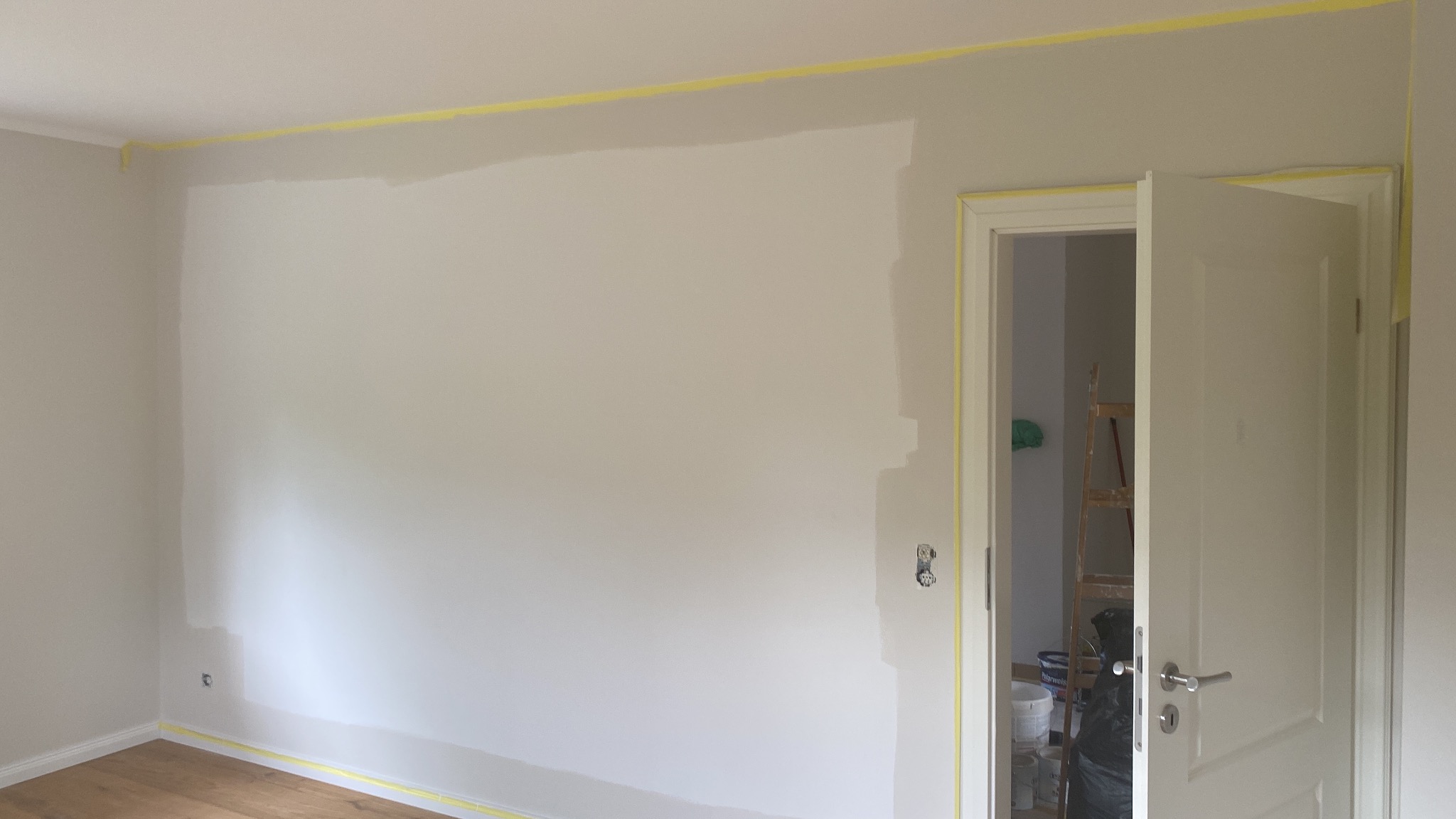 Ściana w trakcie malowania, zabezpieczona taśmą malarską przy suficie, podłodze i wokół ościeżnicy, widoczna drabina i puszki farb w sąsiednim pomieszczeniu.