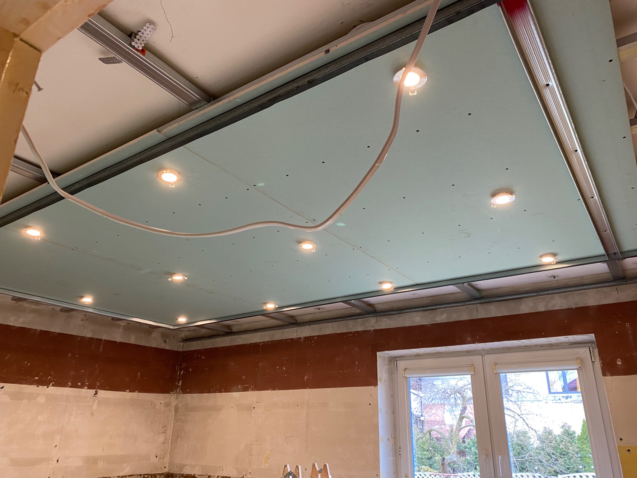 Montaż sufitu podwieszanego z płyt kartonowo-gipsowych z wbudowanym oświetleniem punktowym, widoczne profile metalowe konstrukcji i przewody elektryczne, ściany w trakcie przygotowania do wykończenia.