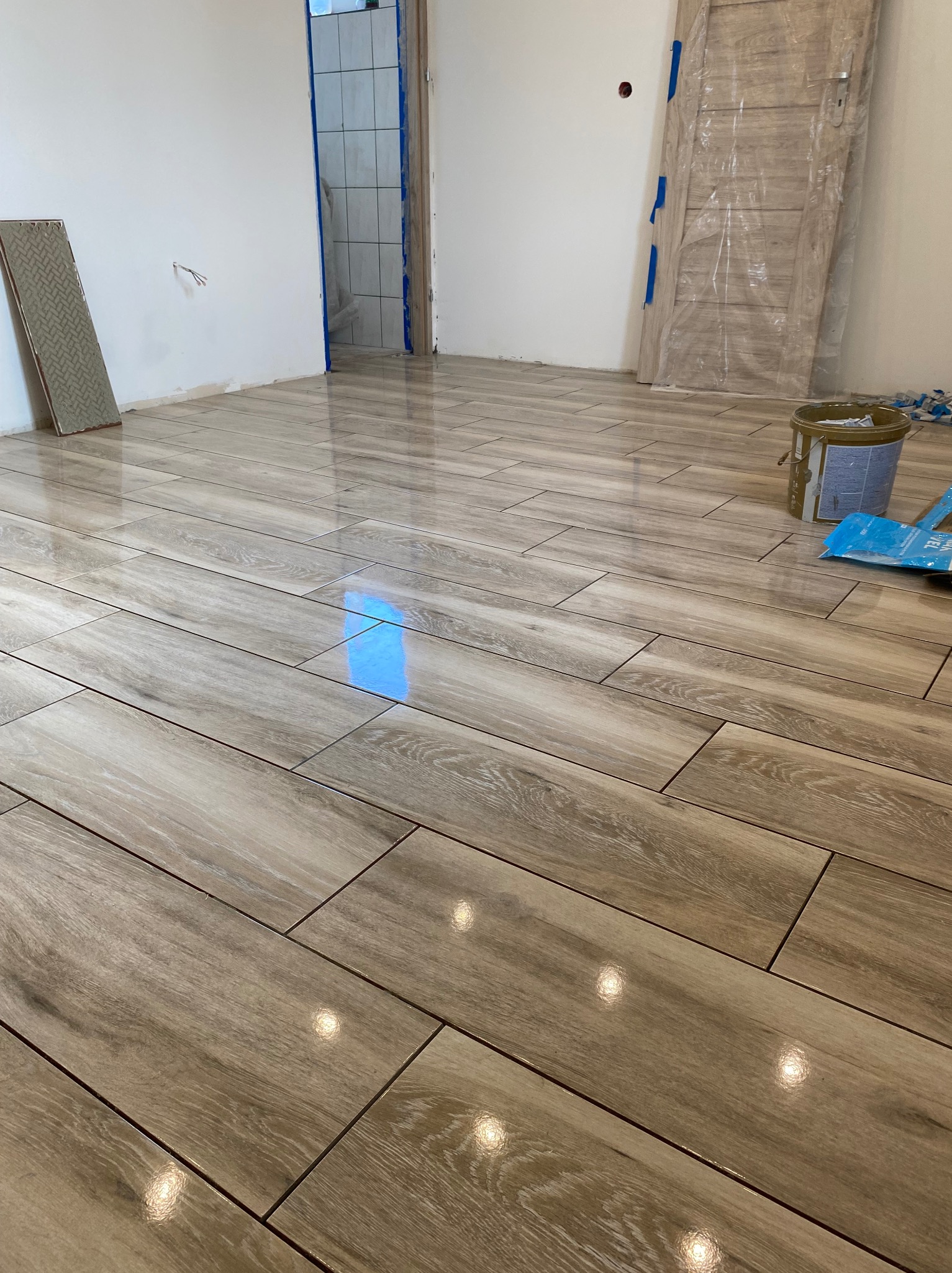 Świeżo ułożona podłoga z paneli imitujących drewno w jasnym odcieniu, widoczne narzędzia i materiały budowlane, fragment zabezpieczonych folią drzwi i płytka podłogowa oparta o ścianę.