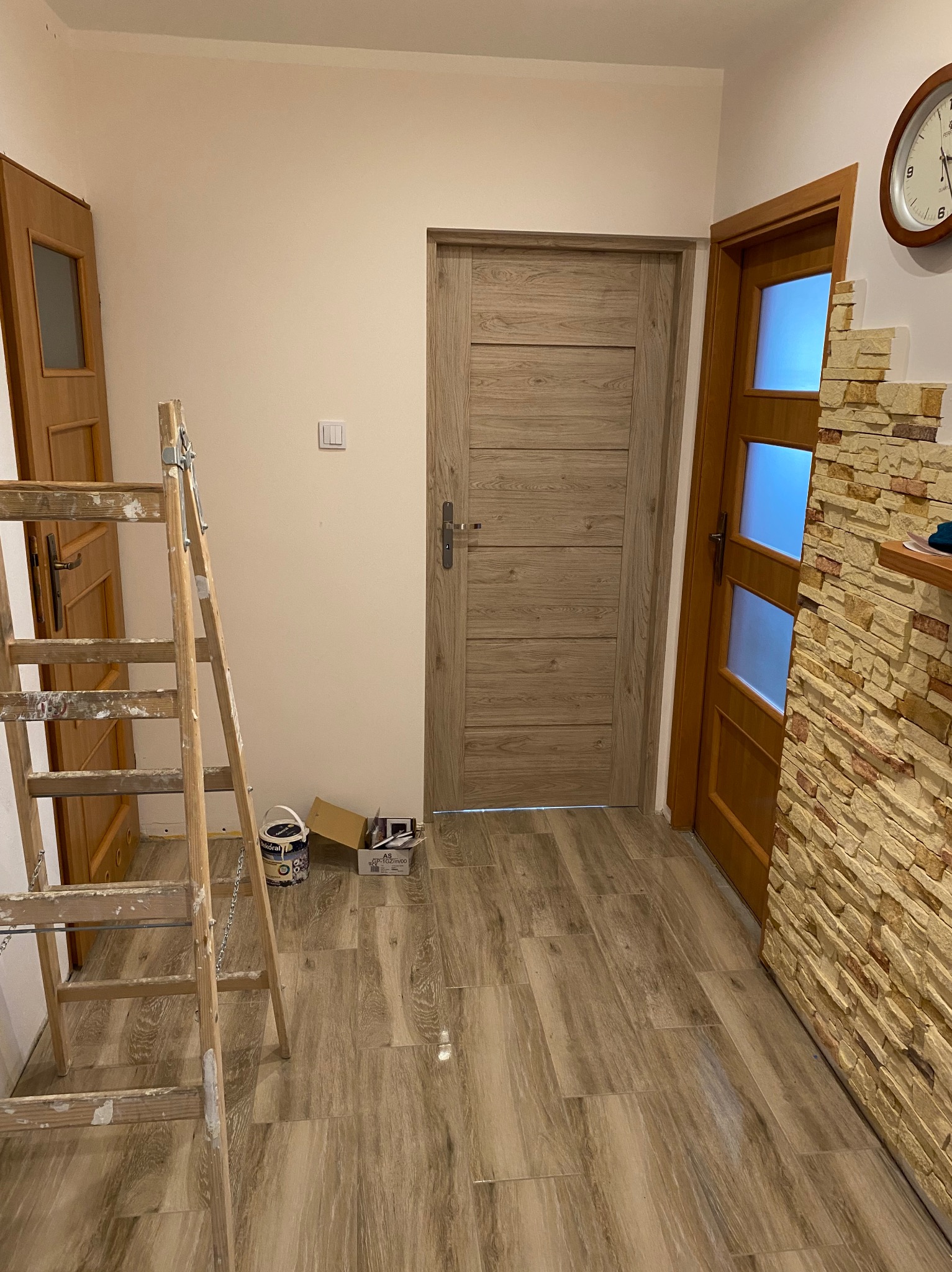 Korytarz w trakcie malowania: drewniana drabina z resztkami farby, puszka farby Dekoral obok ściany przygotowanej do malowania, w tle dwie drewniane drzwi i dekoracyjna ściana z kamienia.