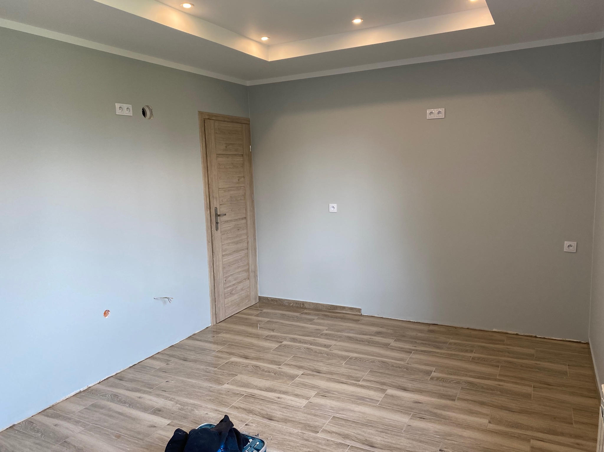 Wnętrze pokoju po remoncie: świeżo położone panele podłogowe imitujące drewno, pomalowane na szaro ściany z widocznymi gniazdkami elektrycznymi, drewniane drzwi i sufit podwieszany z oświetleniem...