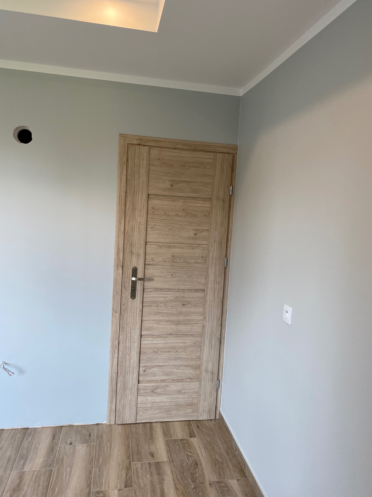 Wnętrze pokoju po remoncie: drewnopodobne panele podłogowe, pomalowane na szaro ściany z widocznym gniazdkiem elektrycznym i otworem na instalację, zamontowane drzwi wewnętrzne z drewnianą okleiną...