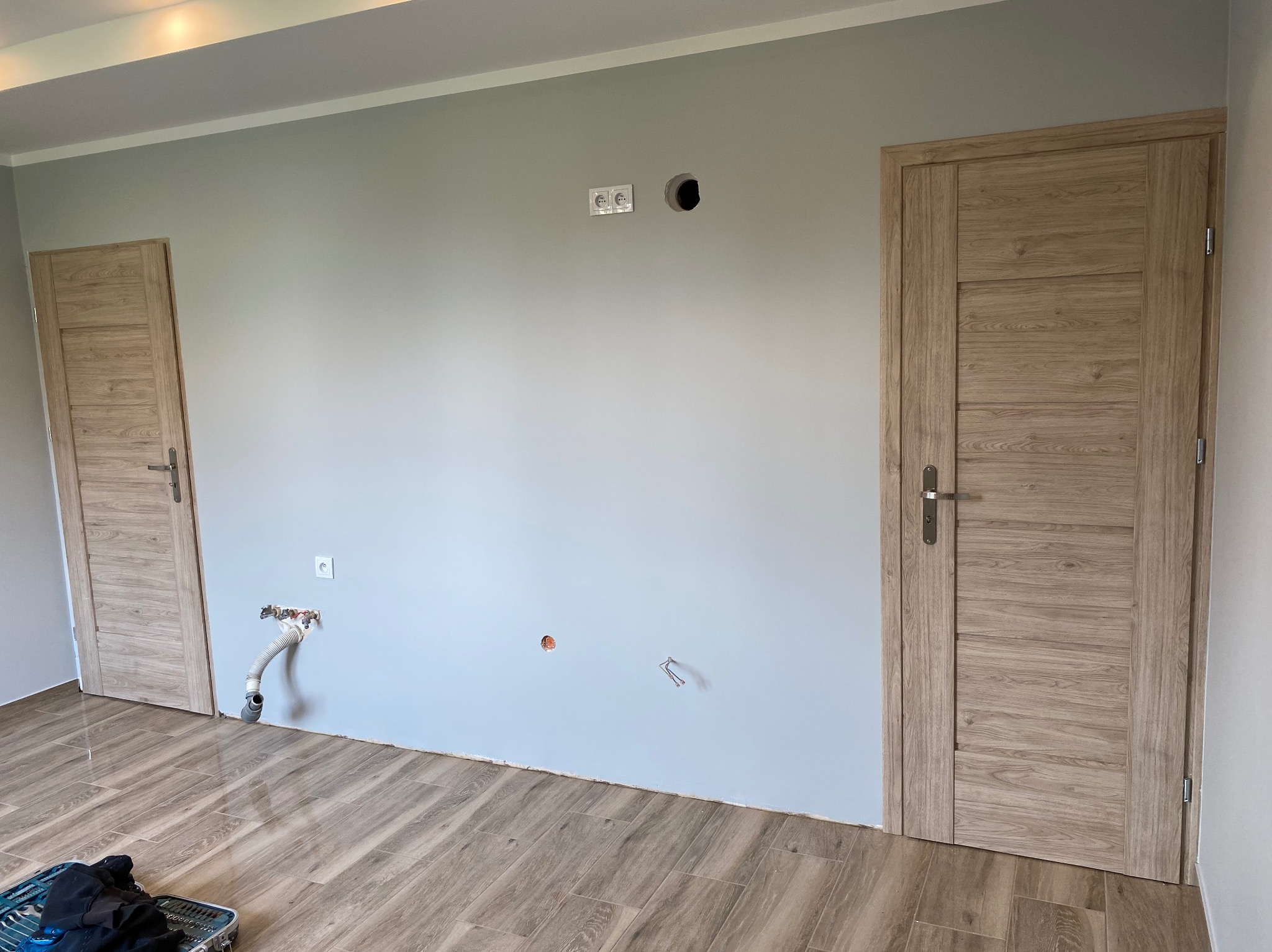 Pokój po odświeżeniu, z dwiema drewnianymi drzwiami, gładkimi, szarymi ścianami z widocznymi gniazdkami elektrycznymi i instalacją hydrauliczną, oraz podłogą wyłożoną panelami imitującymi drewno.