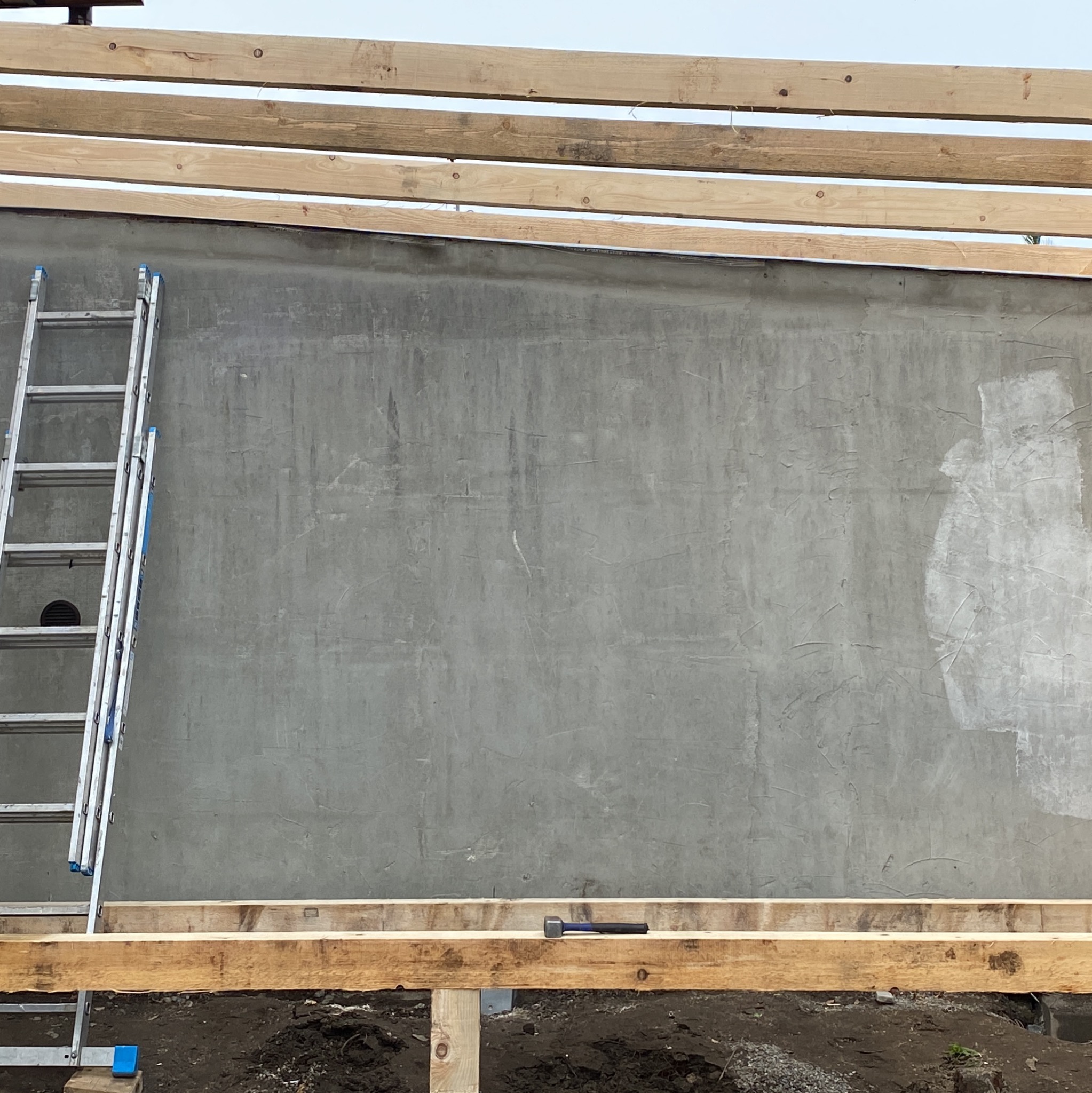 Szara ściana po nałożeniu gładzi, z widocznymi drewnianymi elementami konstrukcji dachu, przystawioną drabiną aluminiową i śladami niedomalowań farbą