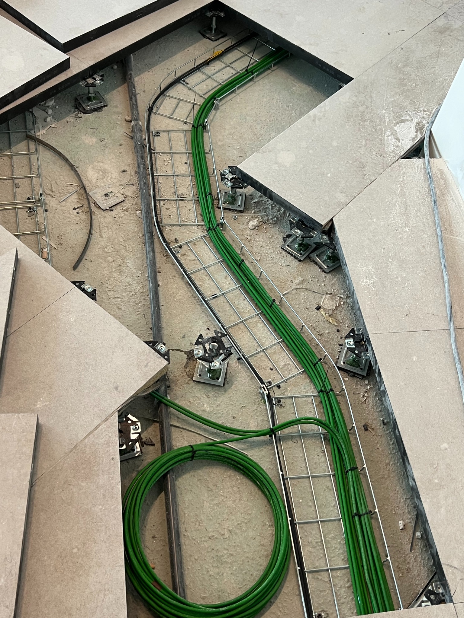Układanie zielonych kabli w metalowej korycie pod podłogą techniczną. Widoczne wsporniki i płyty podłogowe. Etap prac instalacyjnych.