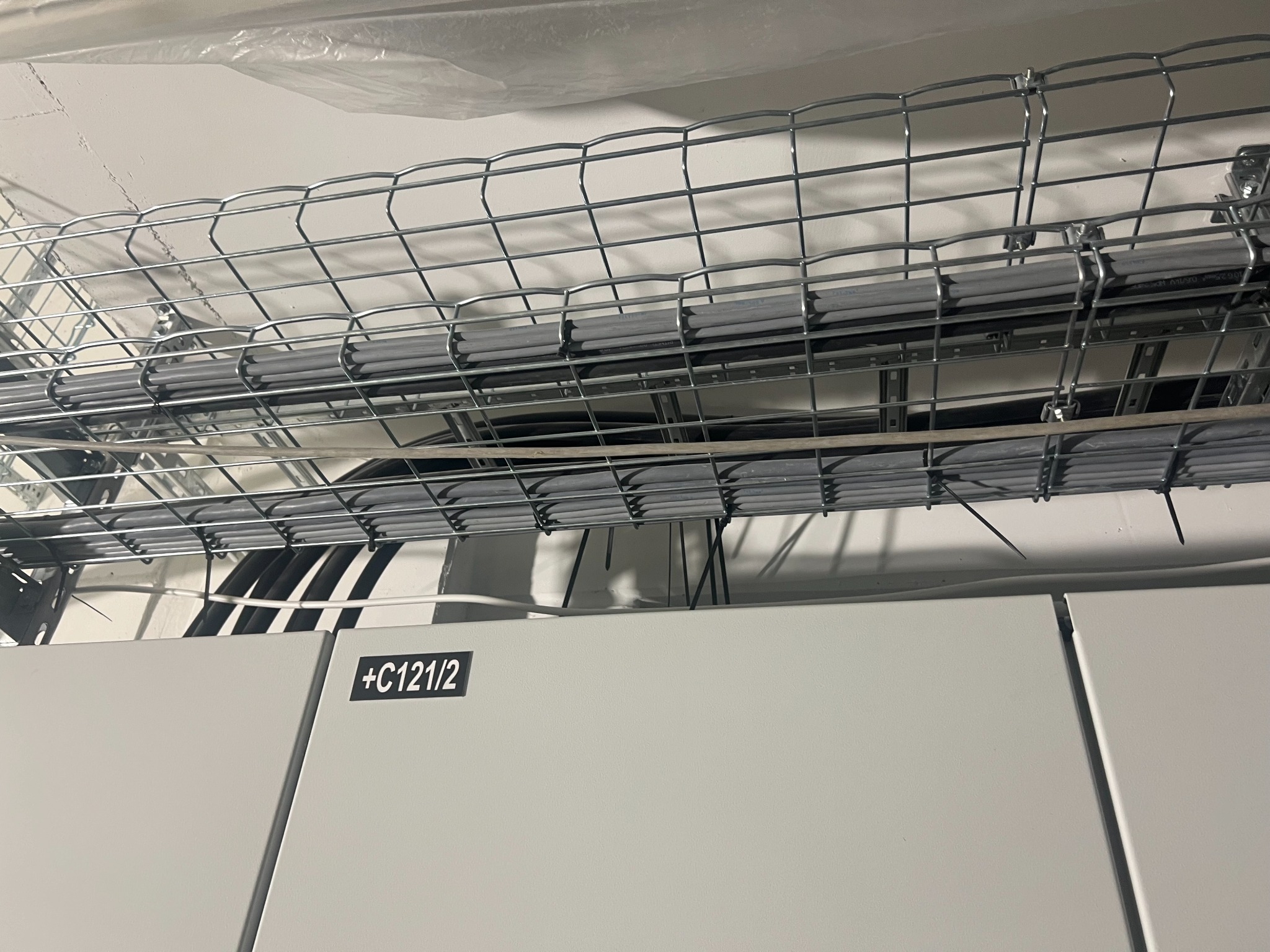 Metalowa korytka kablowa z szarymi rurami i czarnymi kablami zamontowana pod sufitem, widoczna szafa elektryczna z oznaczeniem +C121/2.