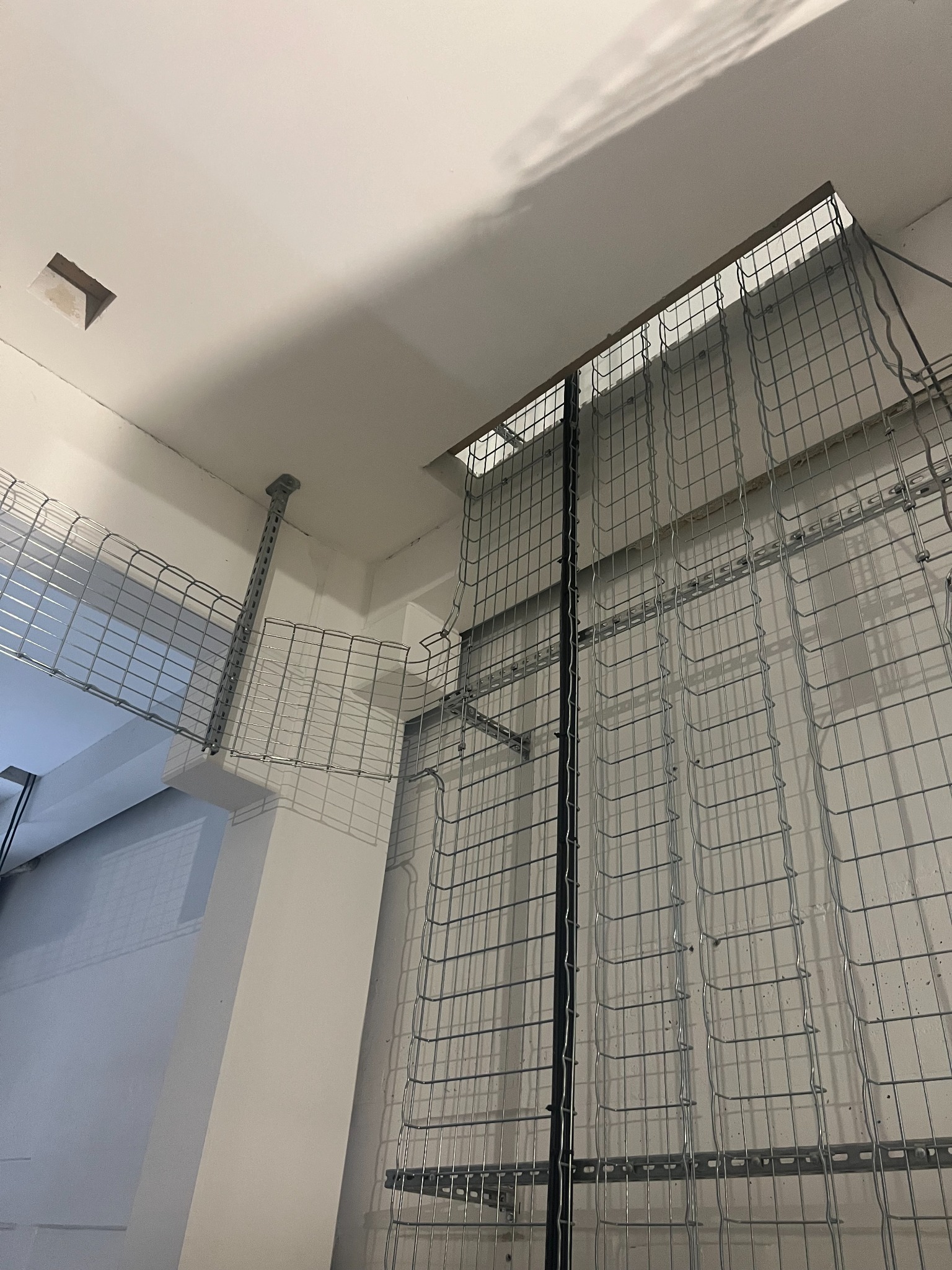 Siatkowe koryta kablowe zamontowane pod sufitem w jasnym pomieszczeniu. Widoczne elementy mocujące i fragment instalacji w narożniku. Ujęcie z dołu.