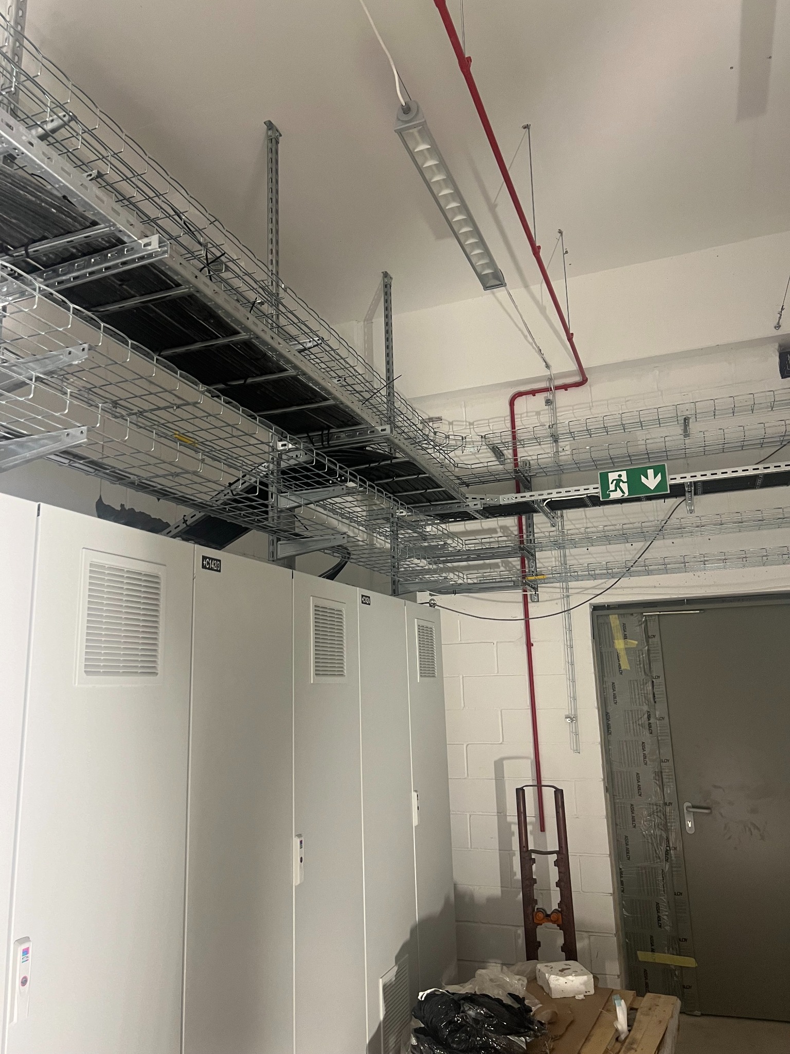 Skomplikowana sieć metalowych korytek kablowych pod sufitem, z widocznymi kablami i rurami. Białe szafy elektryczne w dolnej części kadru. Oznakowanie wyjścia ewakuacyjnego.