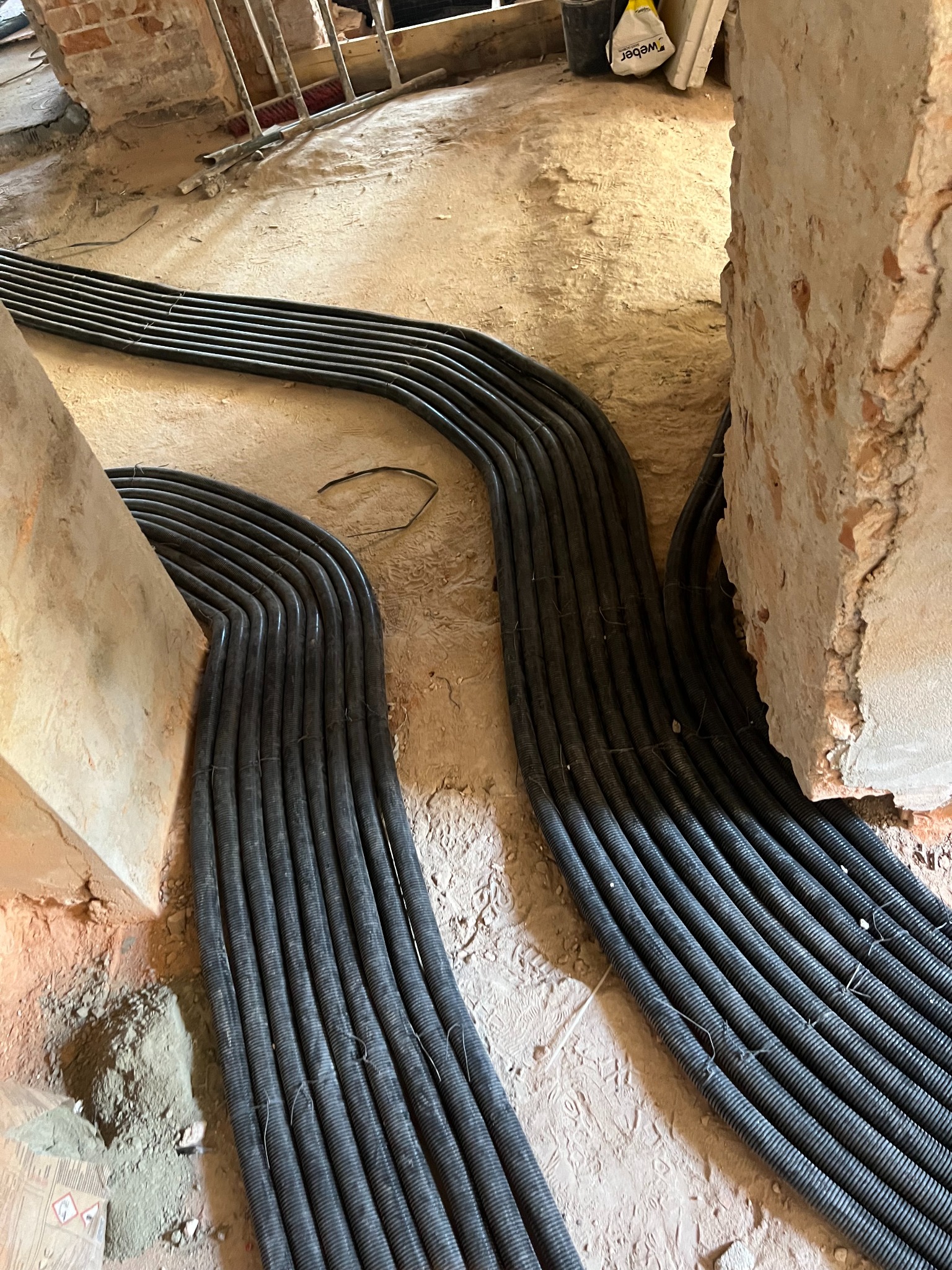 Ułożone w bruzdach podłogi rury instalacyjne w peszlu, prowadzone wzdłuż ścian. Widoczne fragmenty ścian w surowym stanie i podłoże w trakcie prac wykończeniowych.