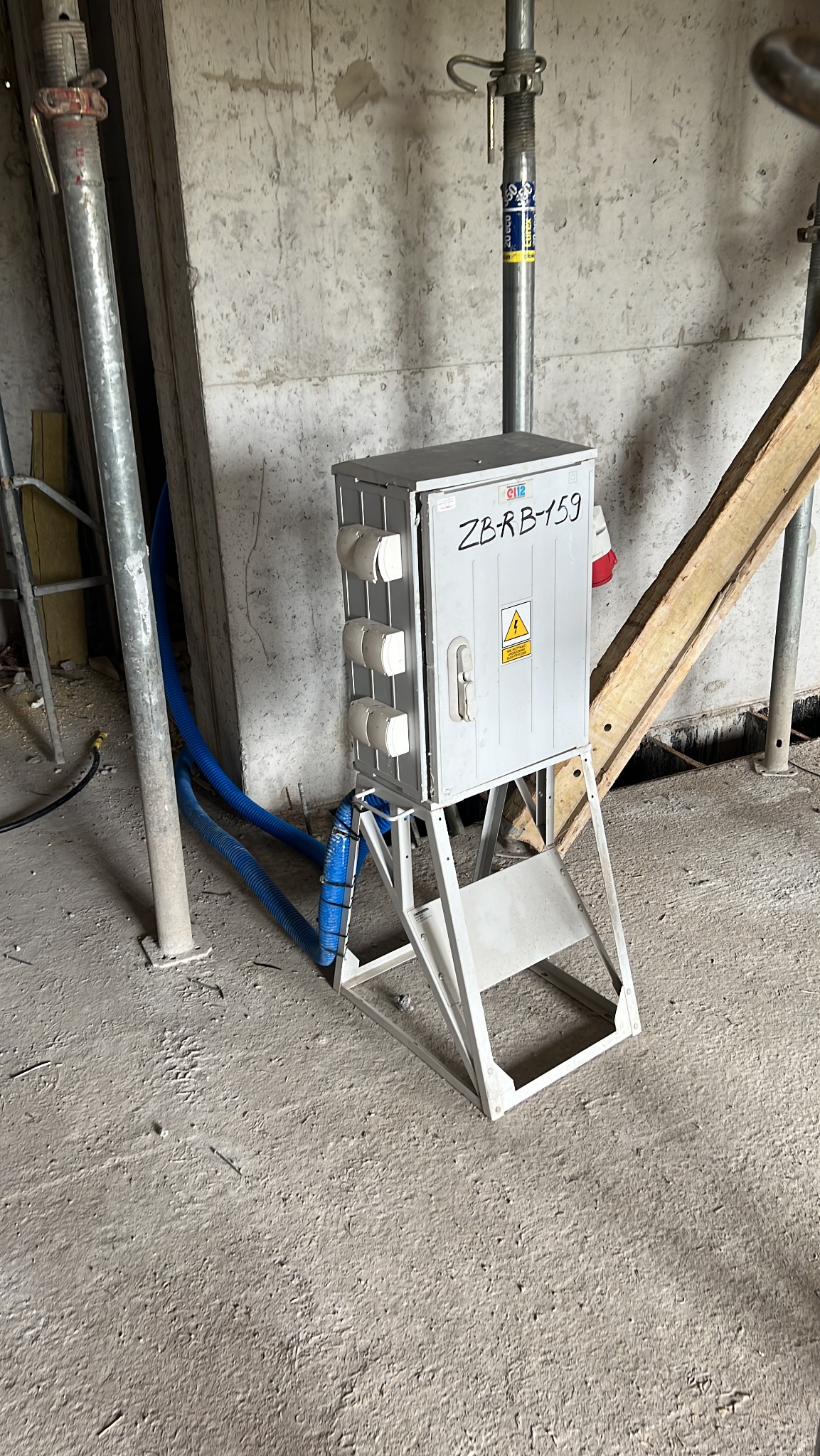 Tymczasowa rozdzielnica elektryczna na budowie, z widocznymi gniazdami i niebieskimi przewodami w peszlu, ustawiona na metalowym stojaku. Szare, surowe tło.