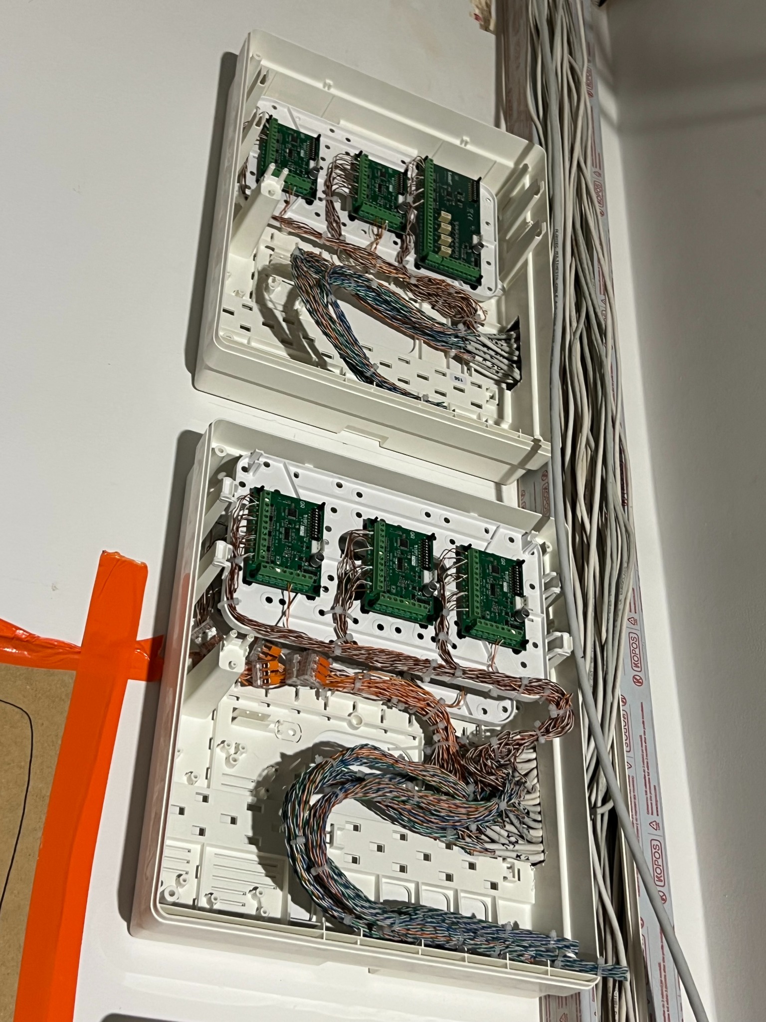 Dwa otwarte panele sterowania z zielonymi płytkami drukowanymi i kolorowymi kablami, zamontowane na białej ścianie, obok wiązka szarych przewodów w listwie.