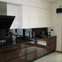arkadia arkadiusz bialobrzewski - Nowoczesna kuchnia z ciemnym blatem i dolnymi szafkami z egzotycznego drewna, górne szafki w kolorze złamanej bieli, czarny panel nad blatem, oświetlenie punktowe i dekoracyjny żyrandol.