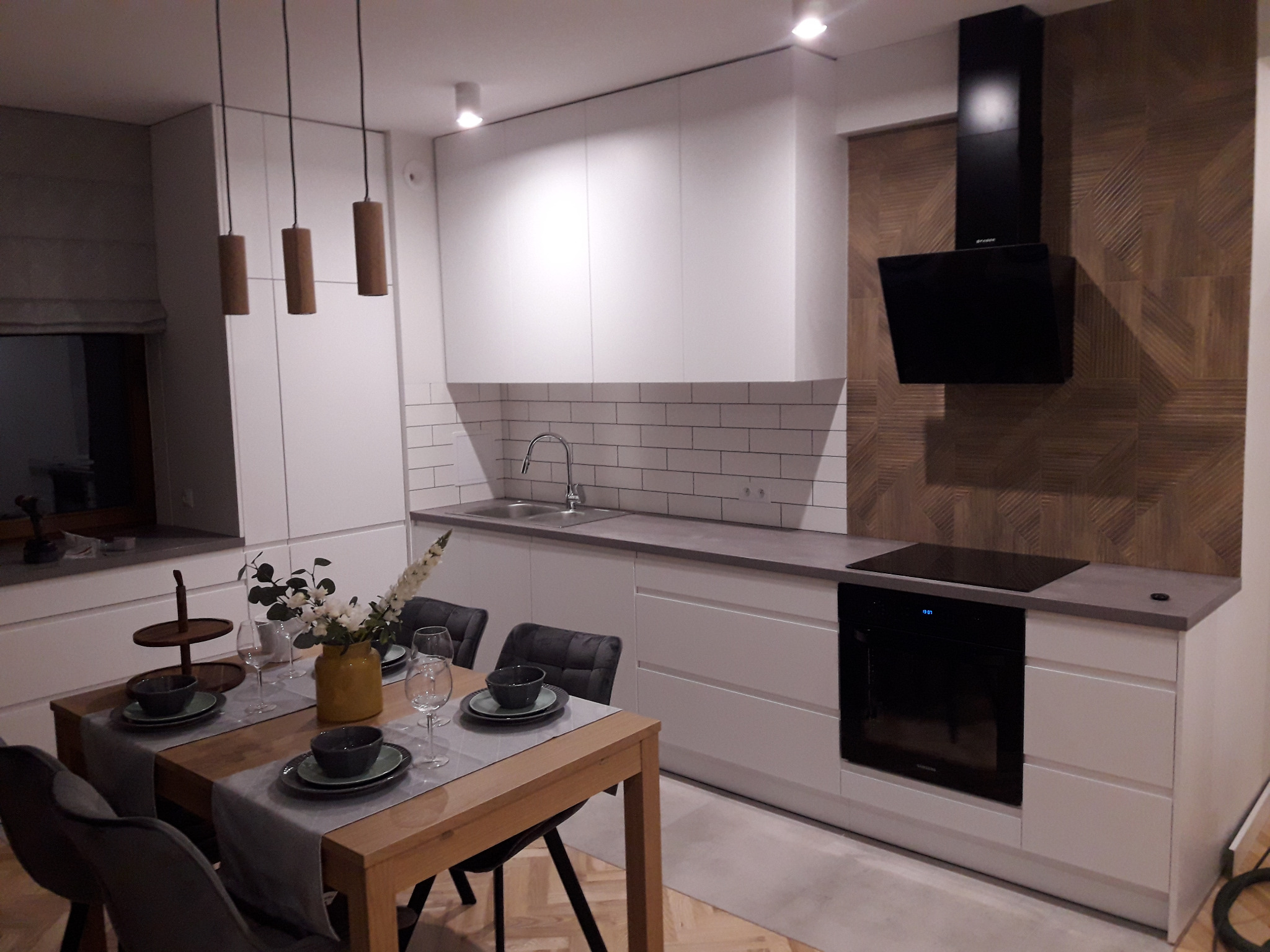 Nowoczesna kuchnia z białymi szafkami, szarym blatem i drewnianym stołem z nakryciem, oświetlona trzema cylindrycznymi lampami wiszącymi.