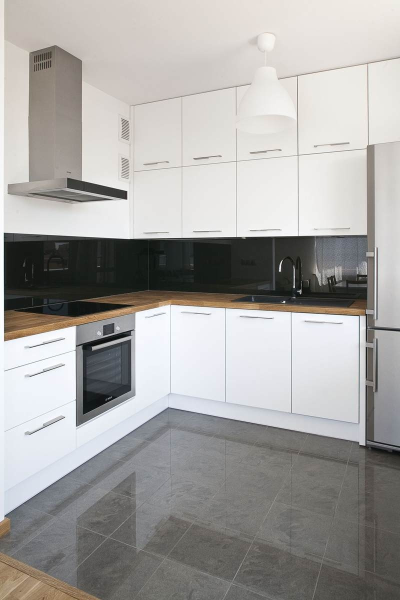 Nowoczesna, jasna kuchnia z białymi szafkami, drewnianym blatem i czarnym panelem szklanym nad nim, uzupełniona stalowym okapem i szarą podłogą z płytek.